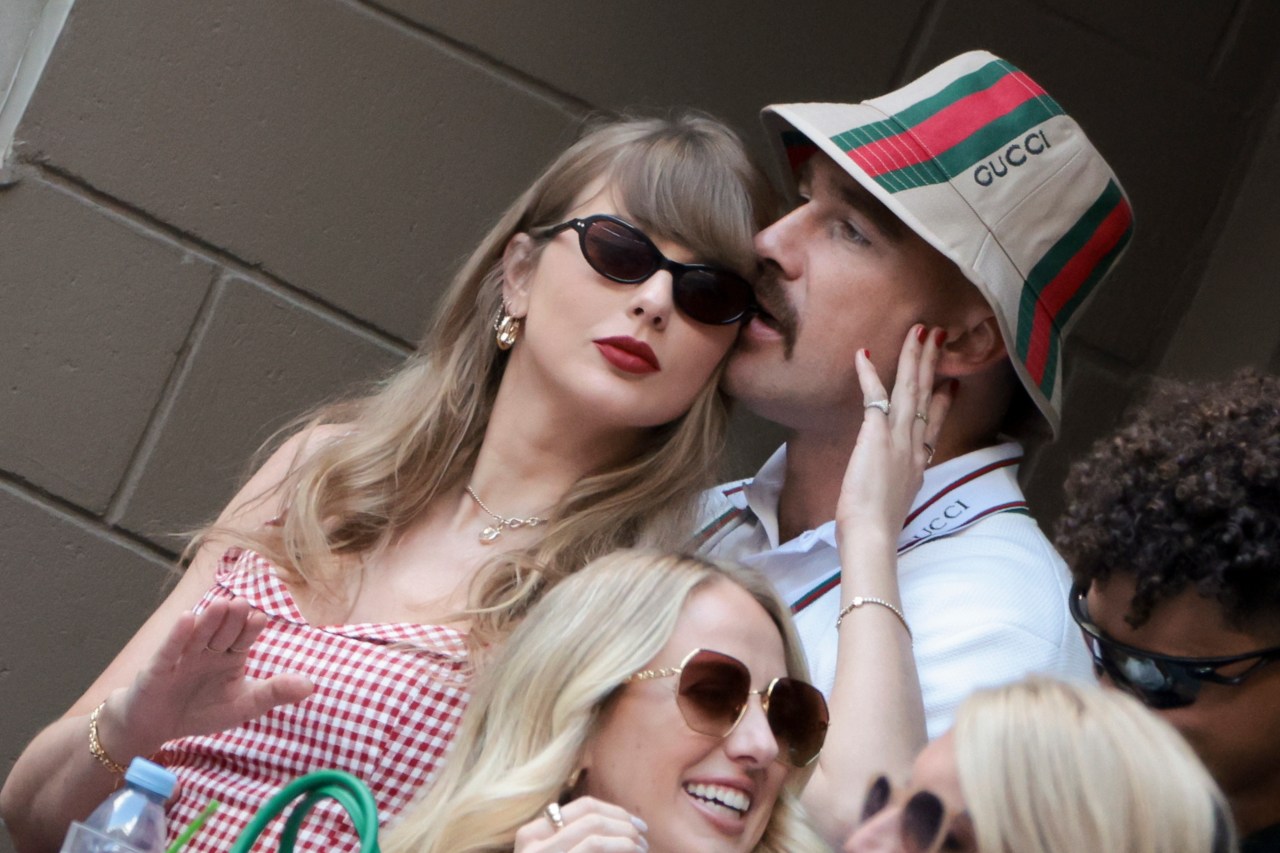 Taylor Swift in sunglasses and a red gingham dress leans close to Travis Kelce, who wears a Gucci bucket hat, in the stands at the US Open.