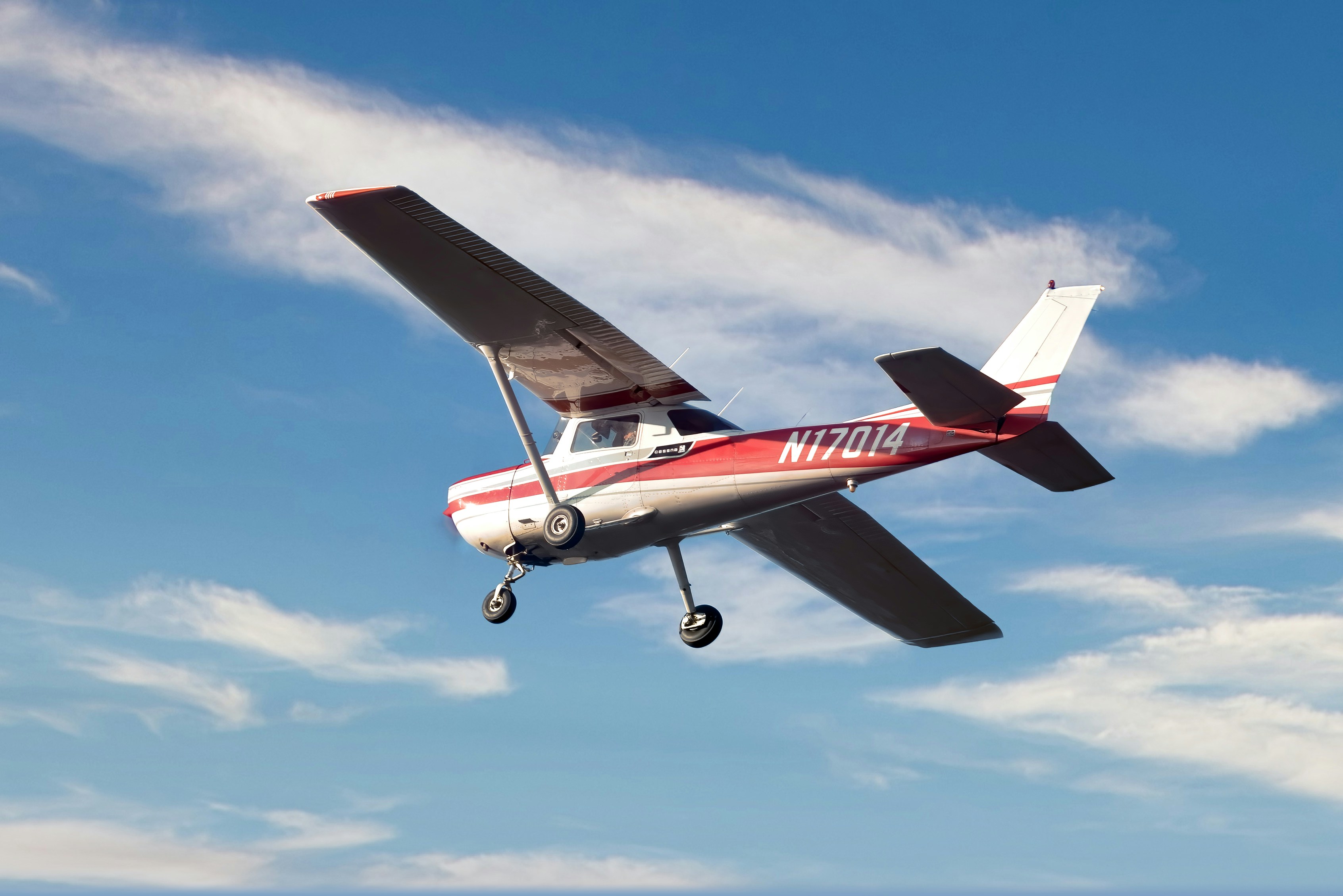 Red and white Cessna single-engine plane with registration N17014 flying against a blue sky with wispy clouds.