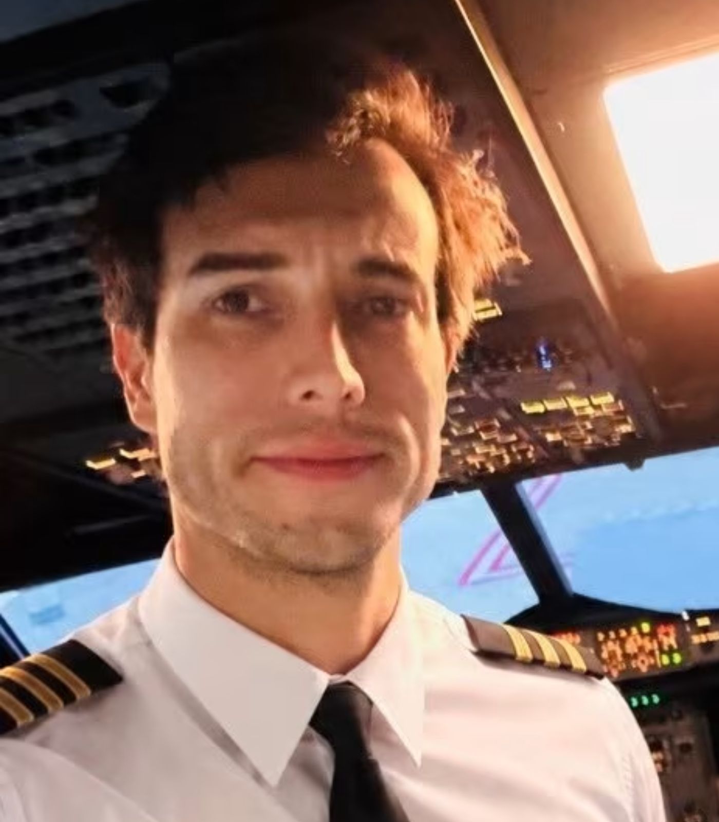Young male pilot in white uniform with gold epaulettes taking a selfie inside an aircraft cockpit.