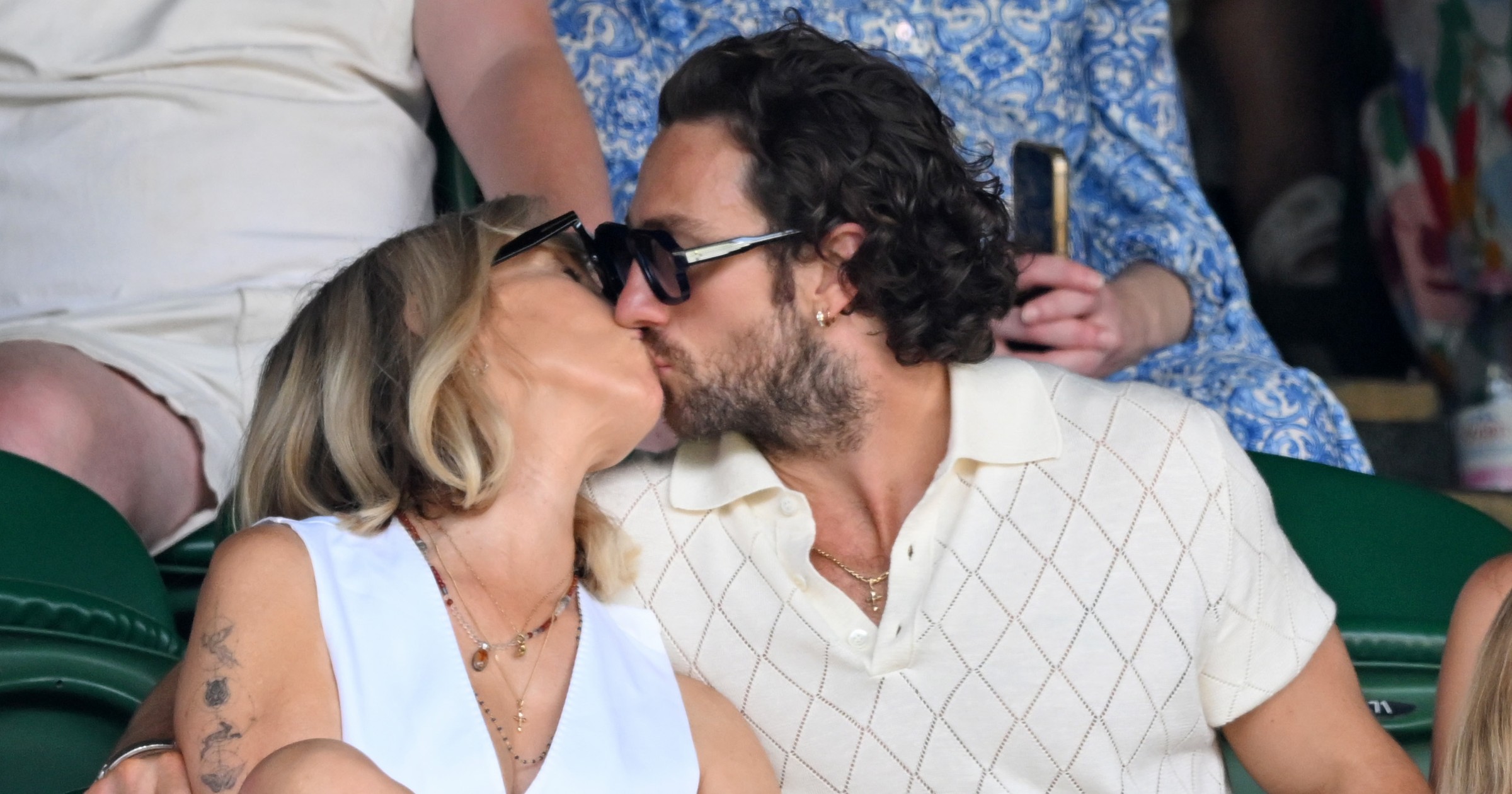 A blonde woman and a curly-haired man, both wearing sunglasses and white outfits, kiss while seated in green stadium stands at Wimbledon.
