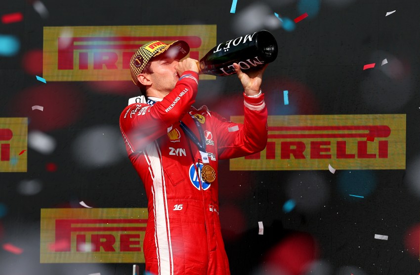 Ferrari driver Charles Leclerc in red race suit drinking from a Moët champagne bottle on the podium surrounded by confetti.