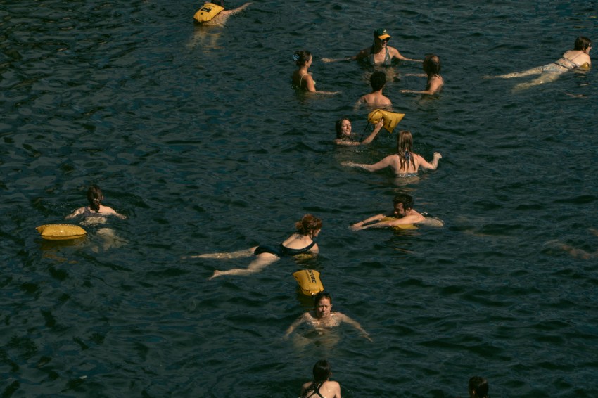 Aerial view of a dozen swimmers in the Seine river, most wearing or holding yellow flotation belts, with dark rippling water surrounding them.
