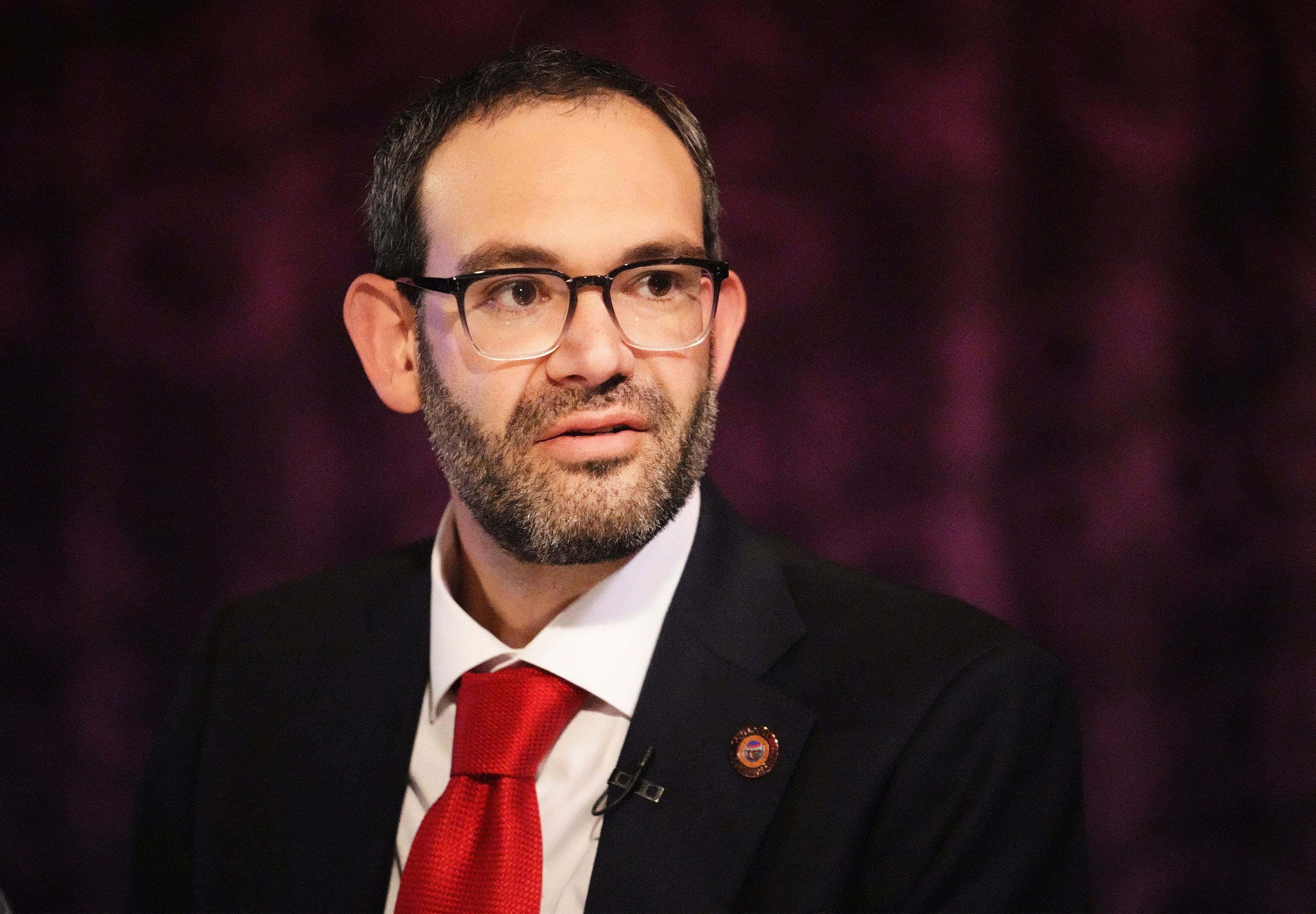 Man with dark hair, beard, and black-rimmed glasses wearing a navy suit and red tie, seated against a purple background.