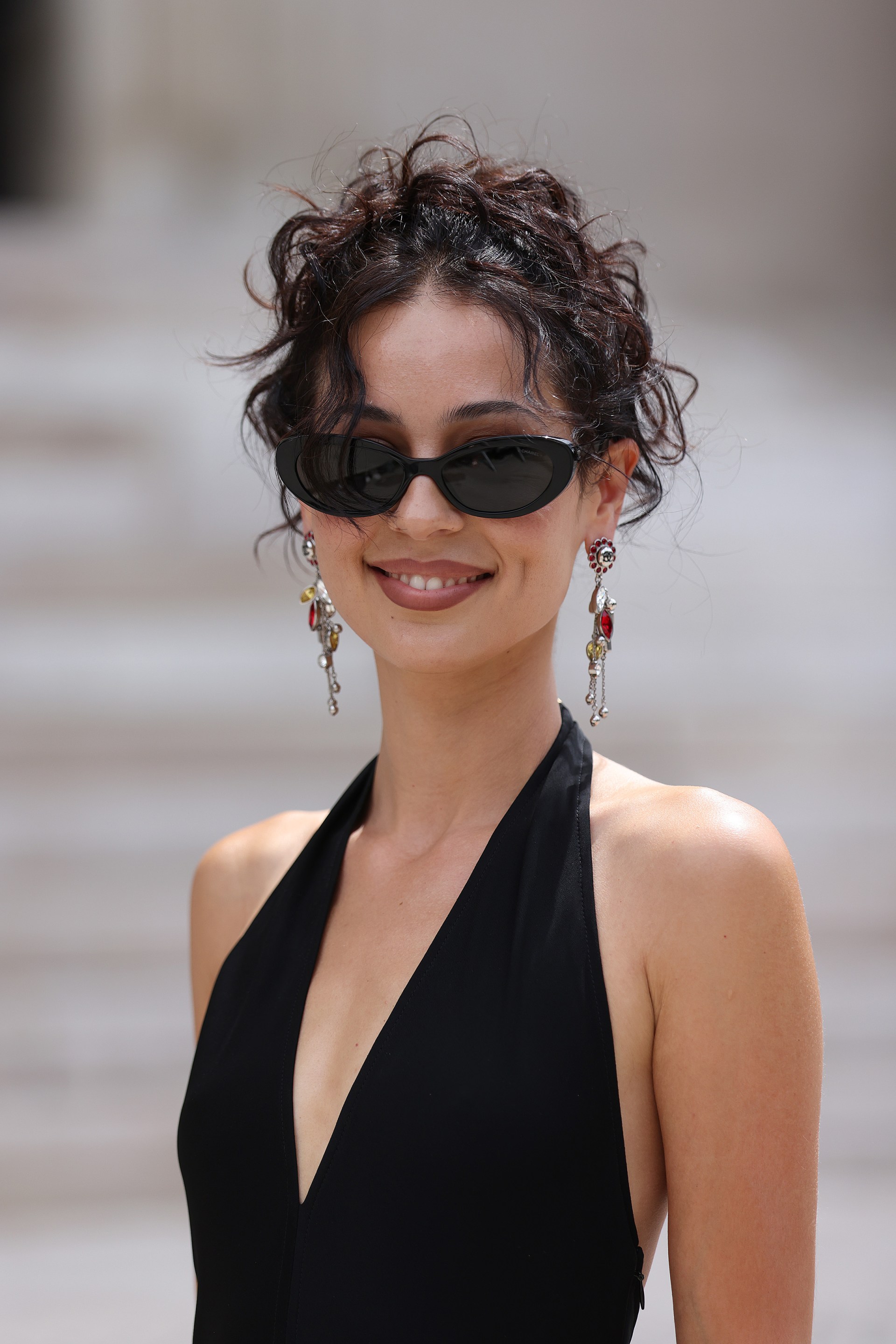 Woman with dark curly updo wearing small oval sunglasses, dangling earrings, and a black halter top, smiling outdoors in Paris.