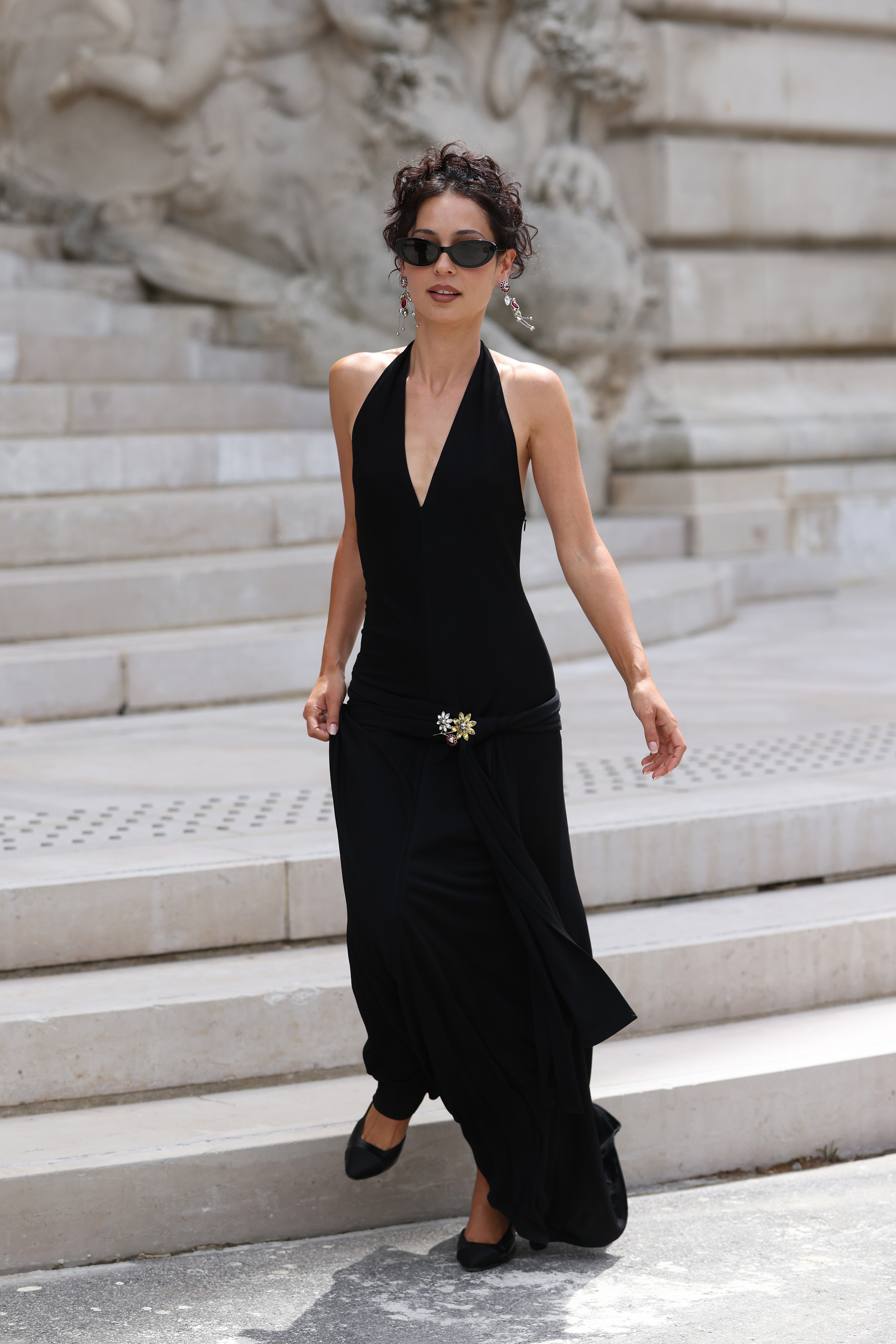 Alexa Demie walking down marble steps in a black halter maxi dress with a jeweled brooch, dark sunglasses, and dangling earrings in Paris.