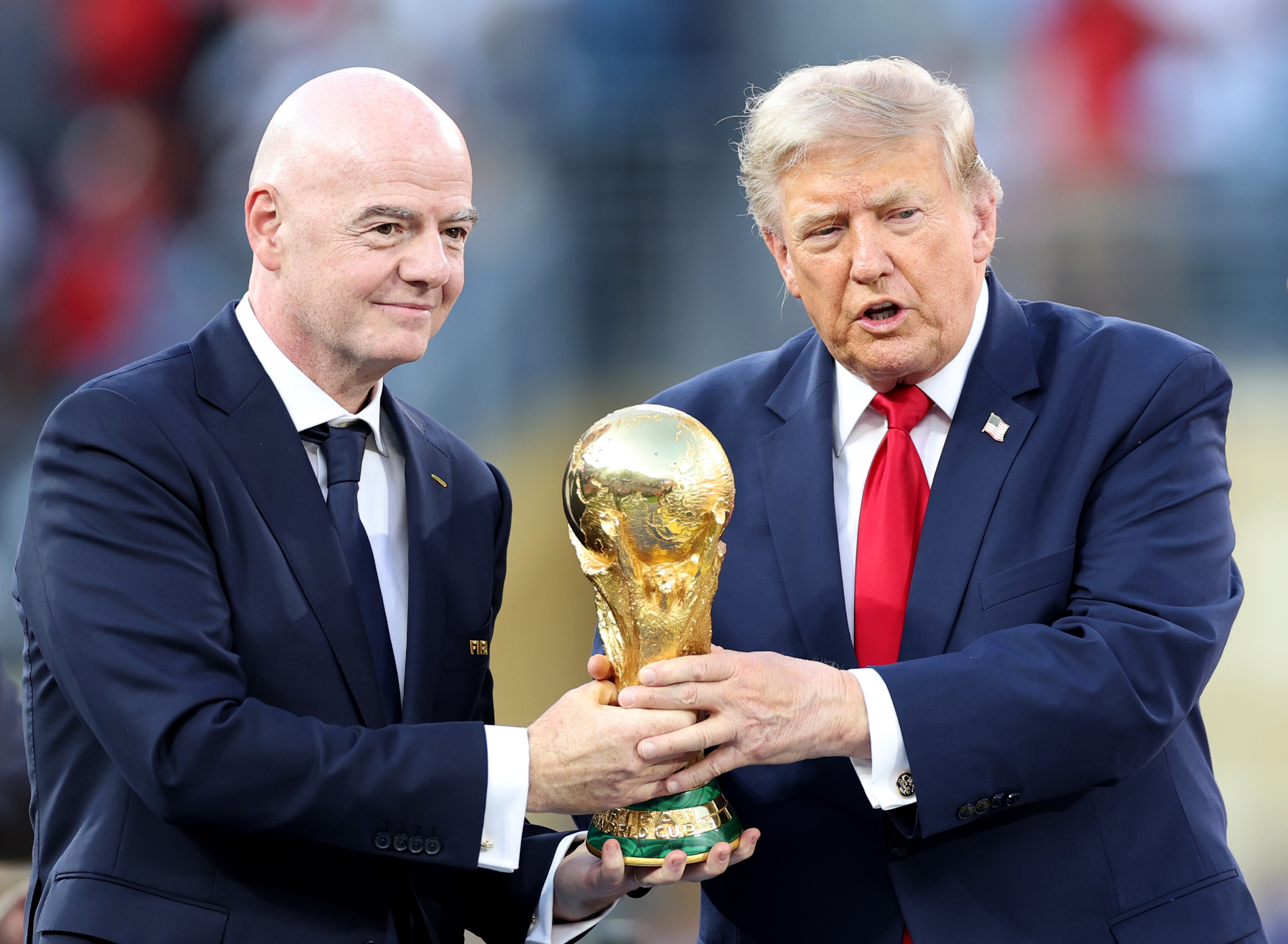 Two men in dark suits jointly holding the gold FIFA World Cup trophy at a stadium ceremony.
