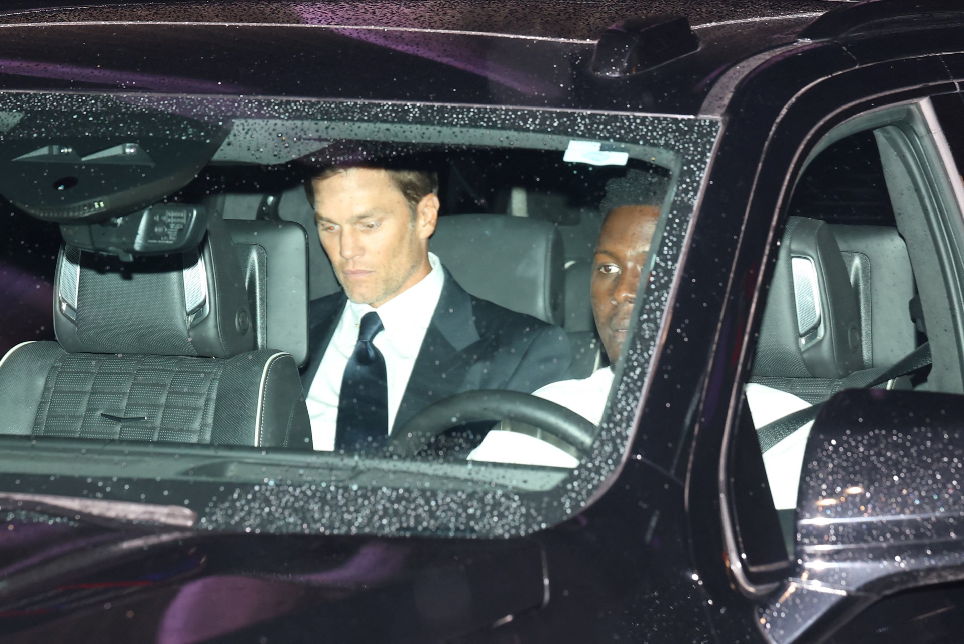 A man in a suit and dark tie sits in the front passenger seat of a rain-speckled SUV at night, with a driver beside him.