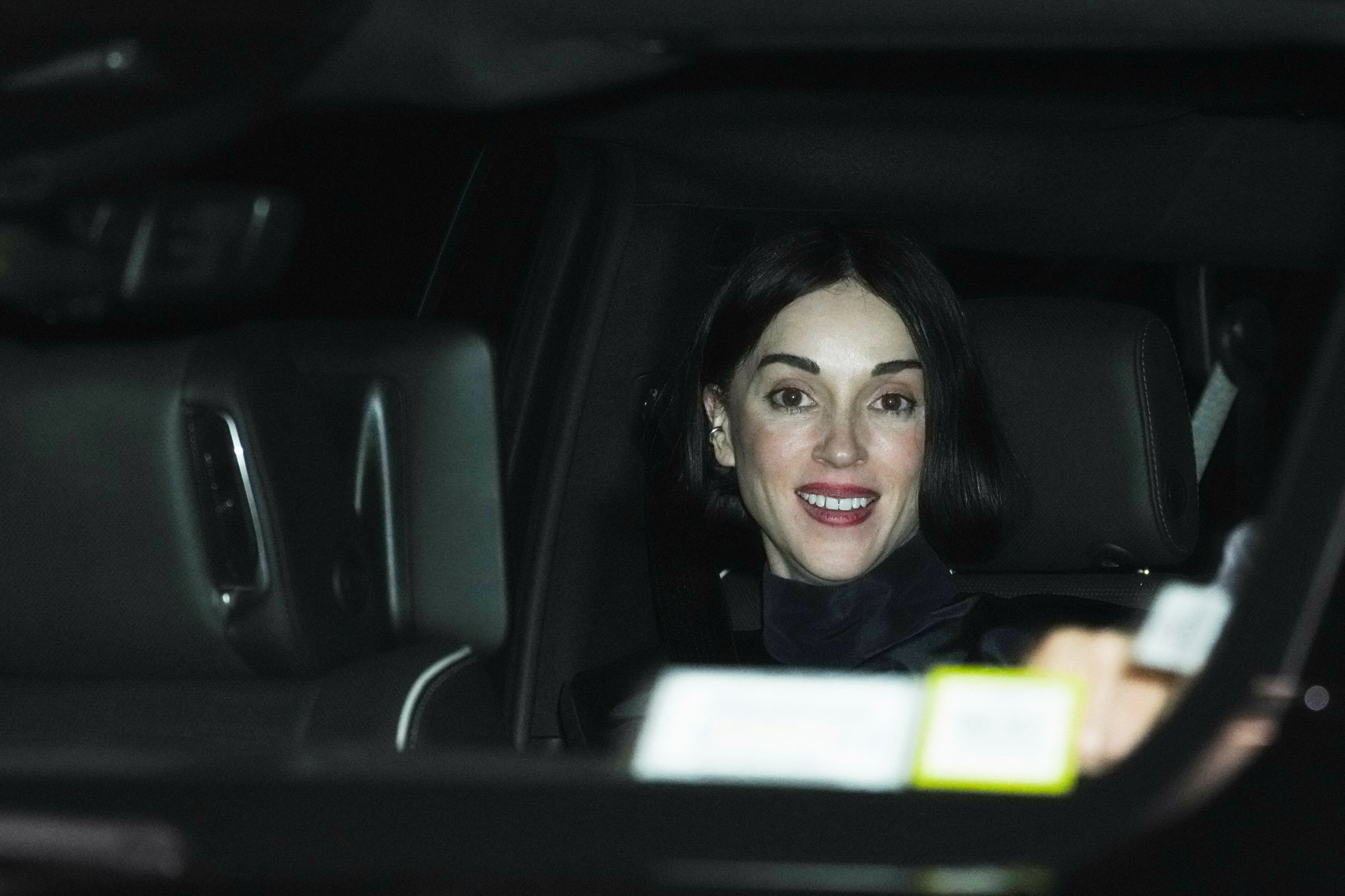 Dark-haired woman with red lipstick smiling from the back seat of a car at night.