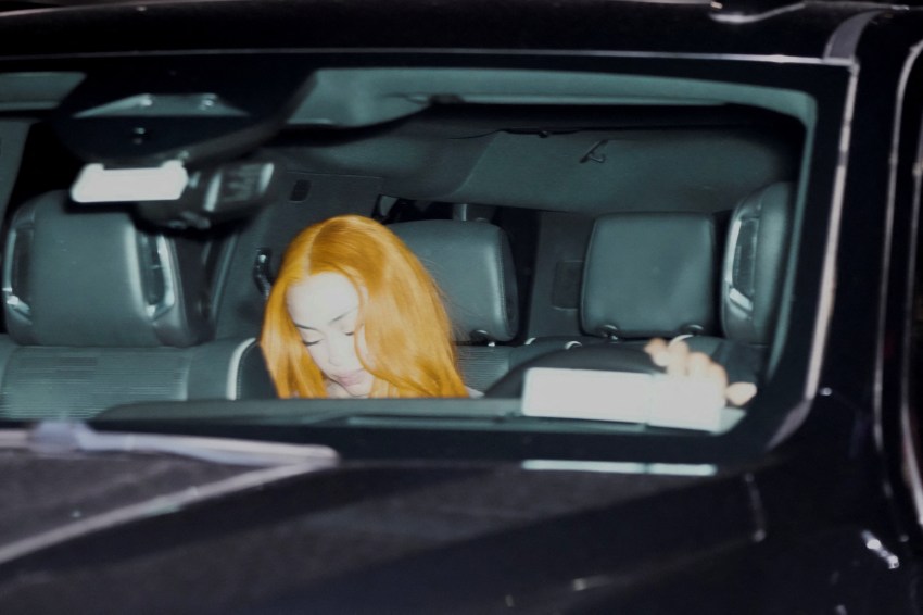A person with long bright orange hair seated in the back of a dark SUV, head bowed, photographed through the vehicle window at night.