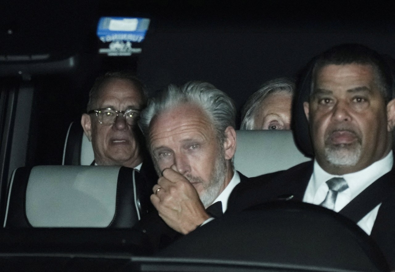 Tom Hanks and Paul McCartney seated in the back of a car at night, with a driver visible in the front seat.