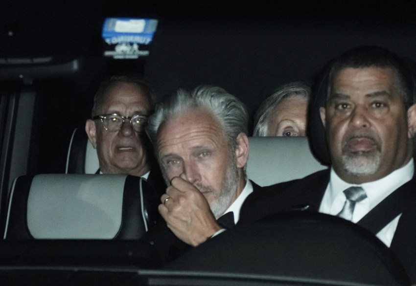 Tom Hanks and Paul McCartney seated in the back of a car at night, with a driver visible in the front seat.