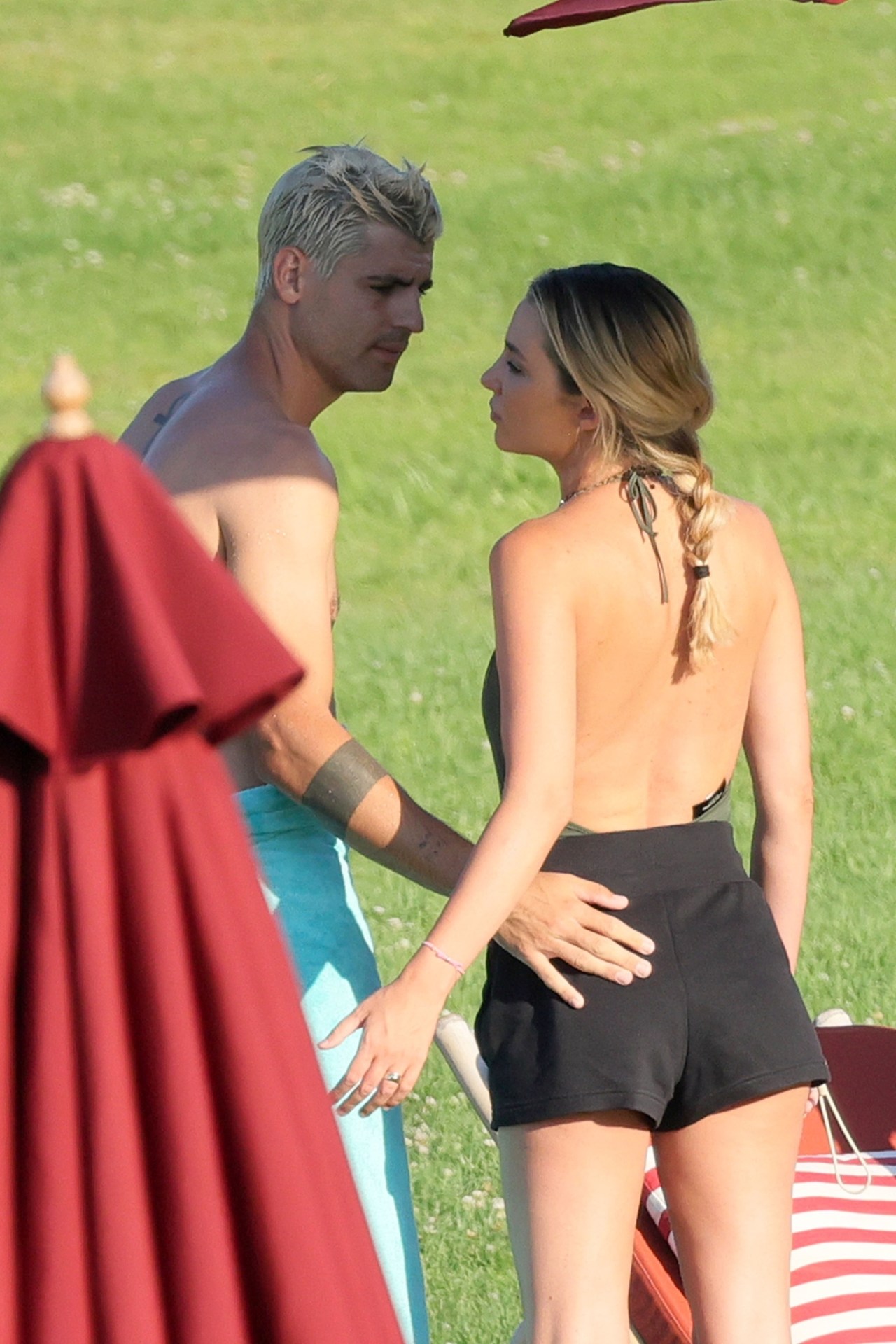 Shirtless man with platinum hair places his hand on a woman's waist as they face each other near sun loungers on a grassy resort lawn.
