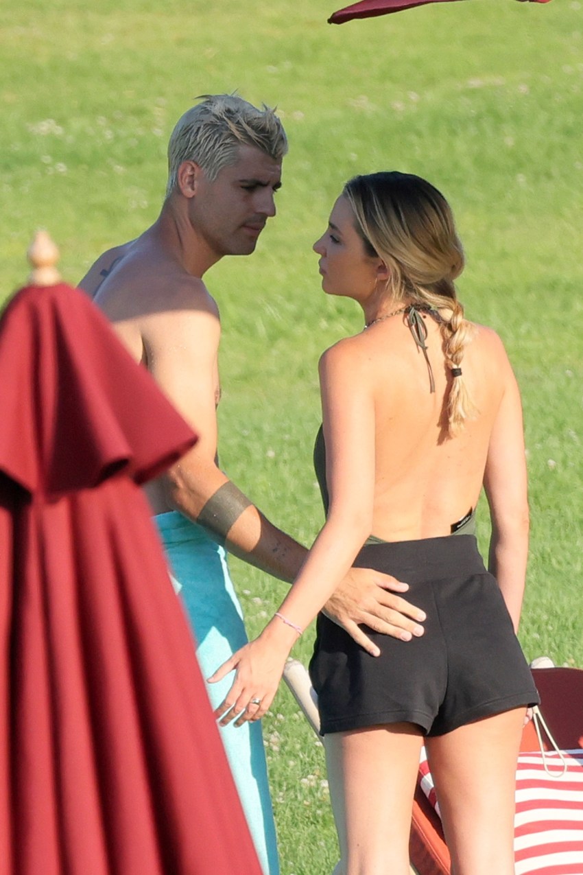 Woman in green halter swimsuit and black Nike skirt talking to a shirtless young boy in swim trunks near poolside bamboo loungers on a grassy lawn.