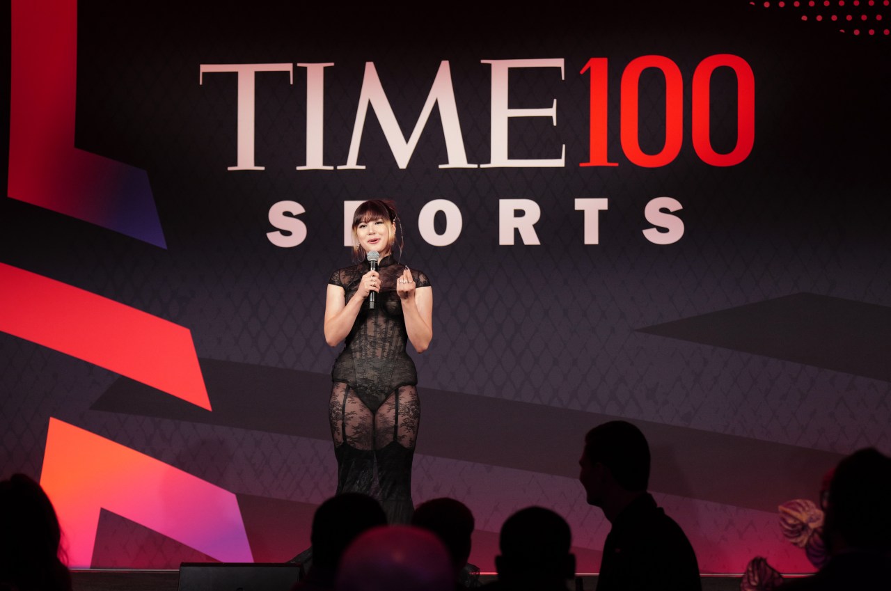 Alysa Liu speaks into a microphone onstage in a sheer black lace cheongsam in front of a large TIME100 Sports backdrop.