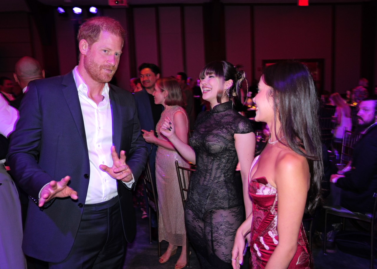 Prince Harry gestures with his hands while talking with Alysa Liu and Madison Chock at an indoor gala event.