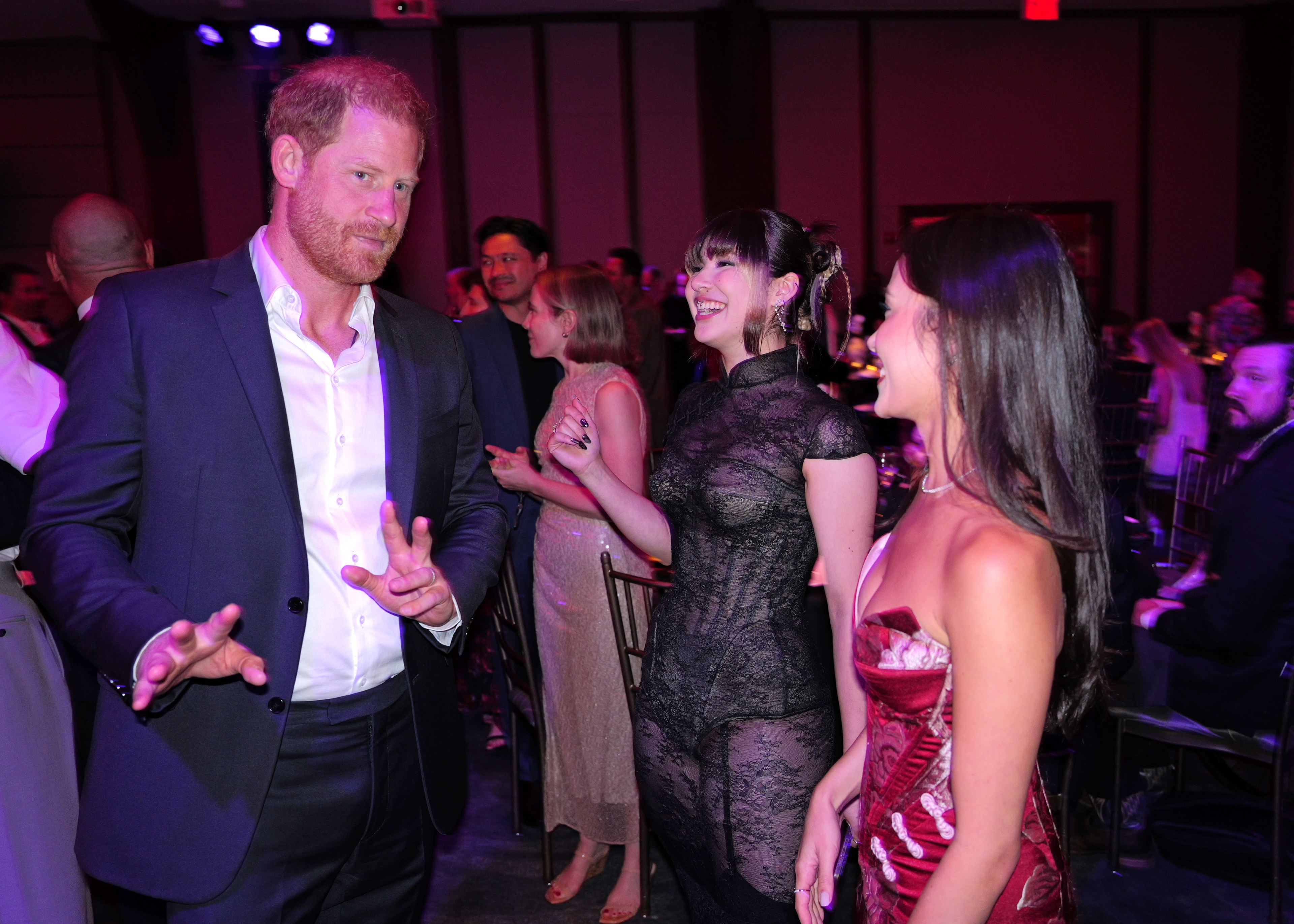Prince Harry gestures with his hands while talking with Alysa Liu and Madison Chock at an indoor gala event.