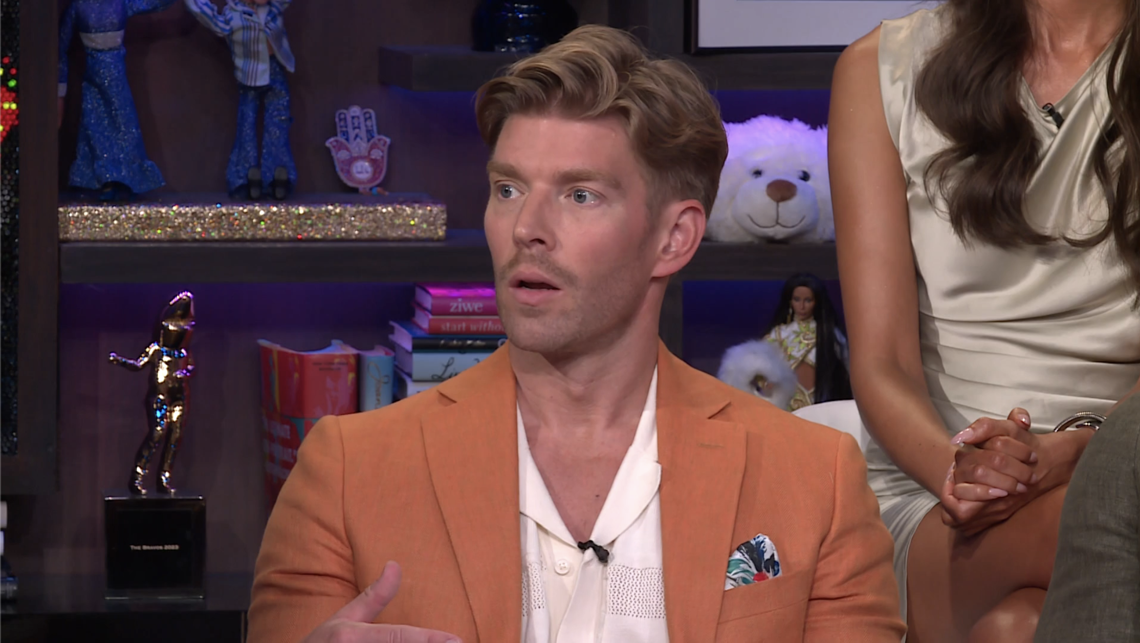 Man in an orange blazer and white shirt with a floral pocket square speaking on a talk show set, with a woman in white partially visible to his right.