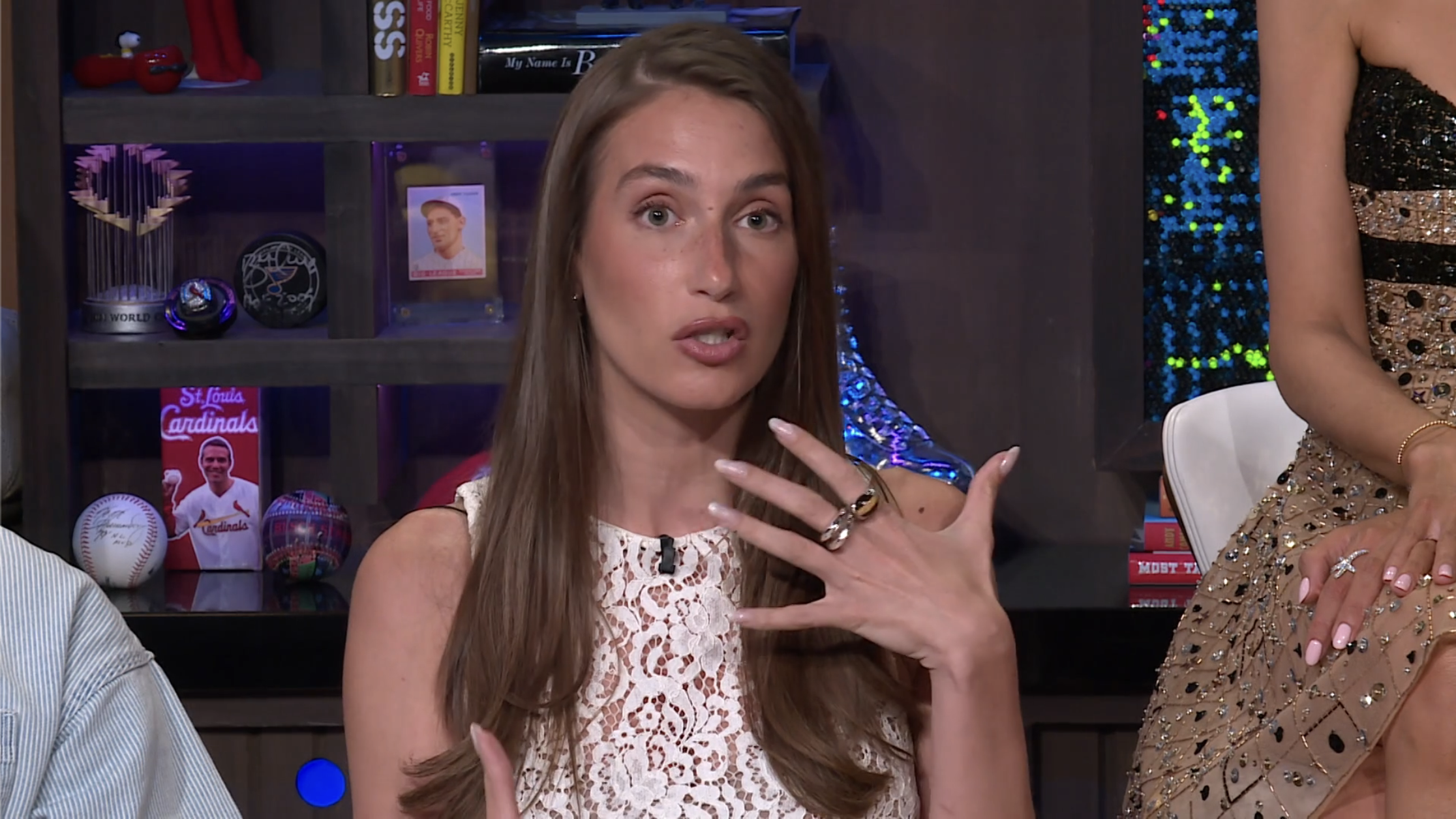 Woman in a white lace sleeveless dress gesturing with her hand while speaking on a talk show set with bookshelves and sports memorabilia in the background.