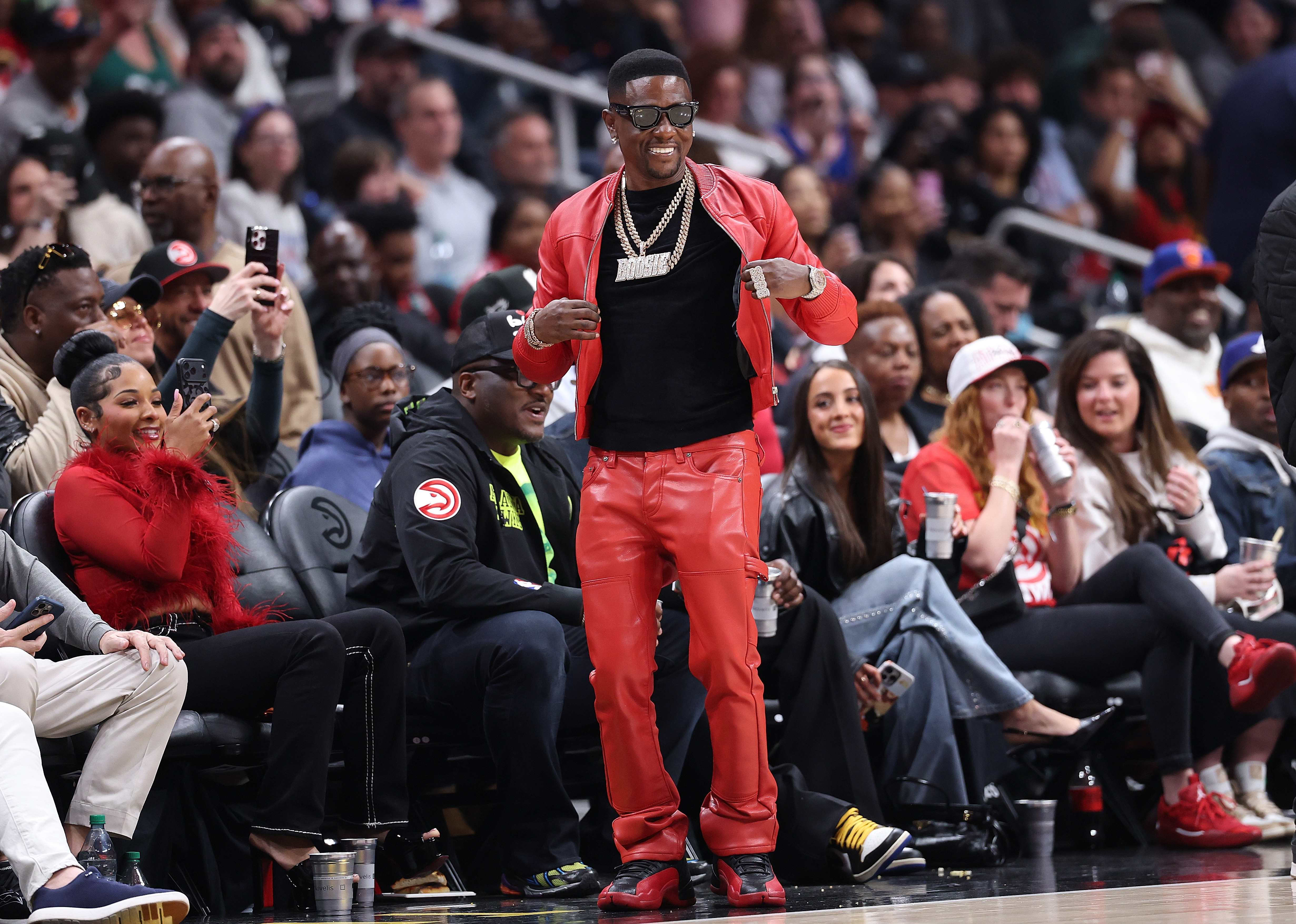 Rapper Boosie Badazz in a red leather jacket and pants dancing courtside at an Atlanta Hawks NBA game, surrounded by fans in the stands.