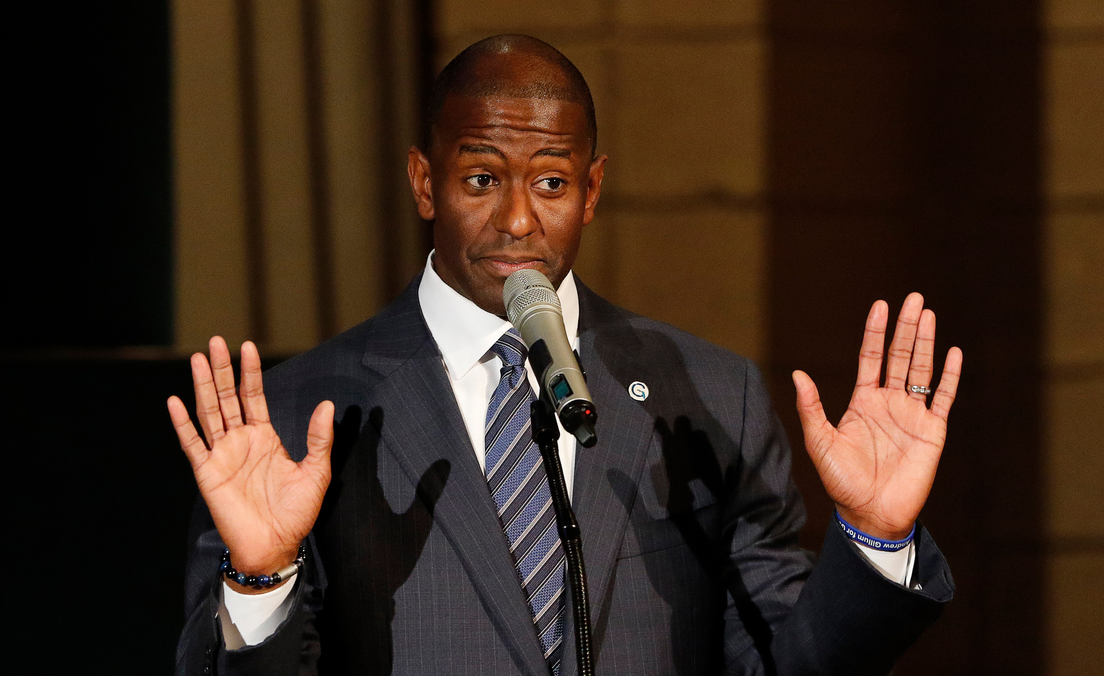 Andrew Gillum in a dark suit speaks at a microphone with both hands raised, palms facing outward, at a church event in Fort Lauderdale, Florida.