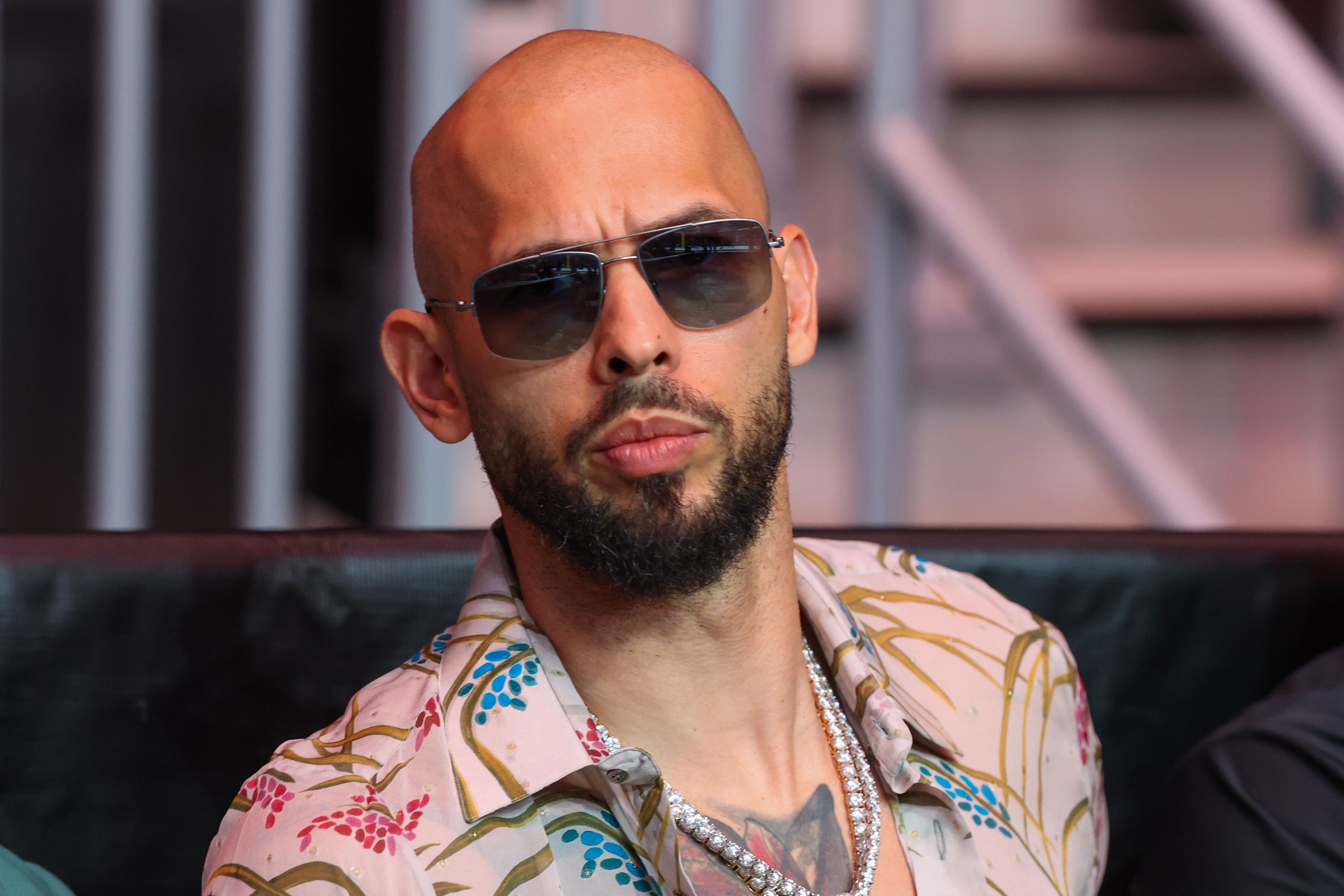 Bald man with beard and sunglasses wearing a floral pink shirt and diamond chain necklace seated at an event.