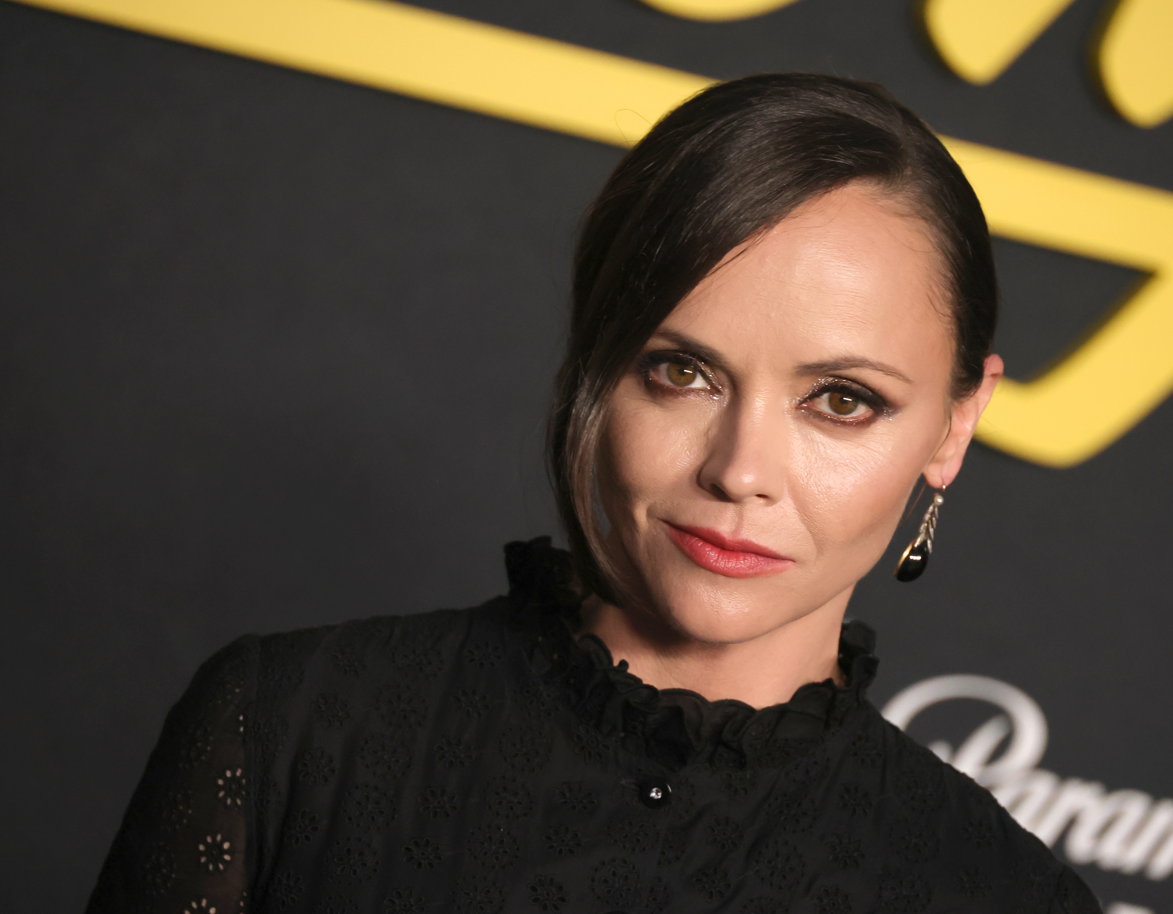 Woman with dark hair pulled back wearing a black eyelet lace dress with ruffle neckline at a red carpet event.