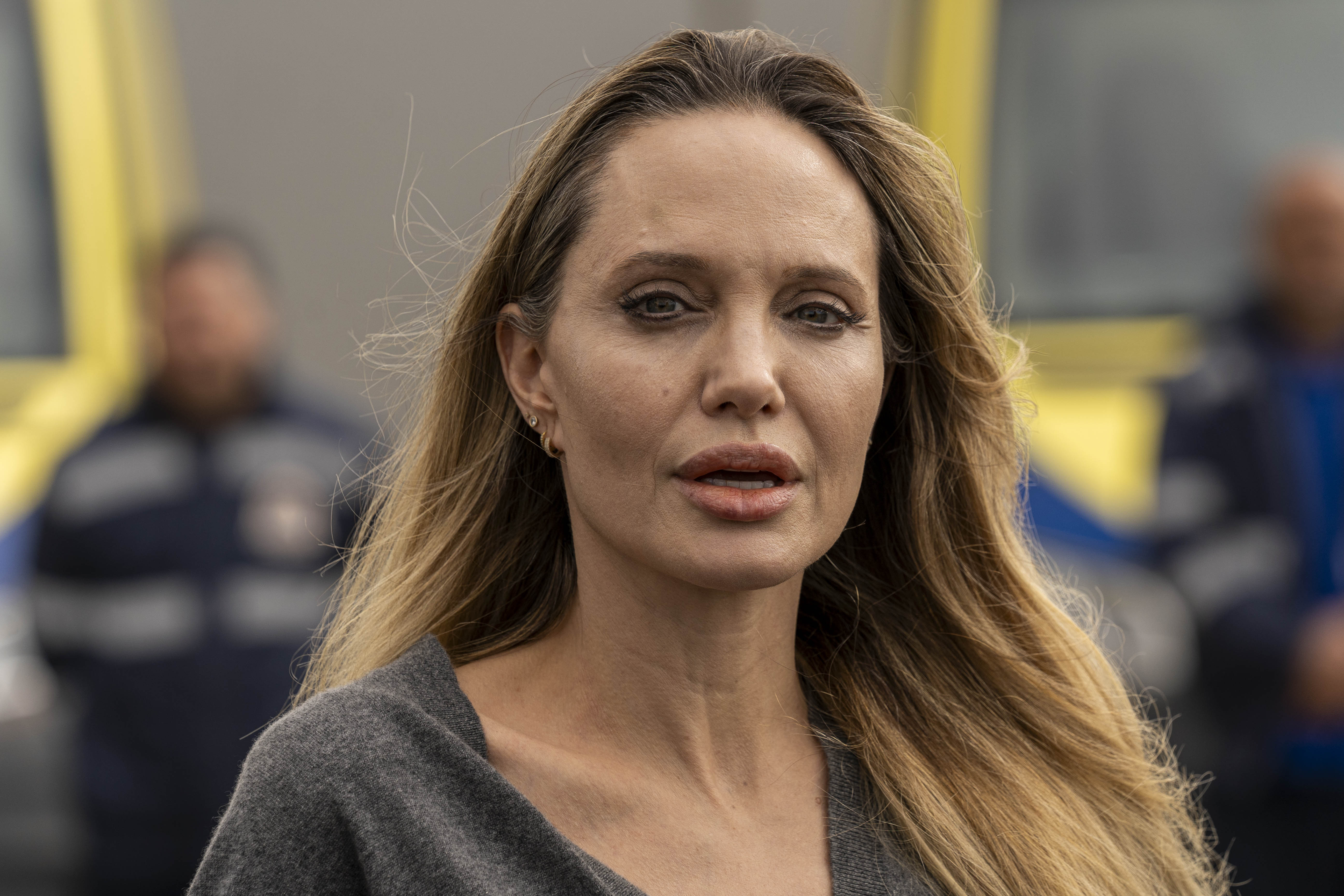 Close-up of a woman with long blonde hair speaking outdoors, uniformed personnel visible and blurred in the background.