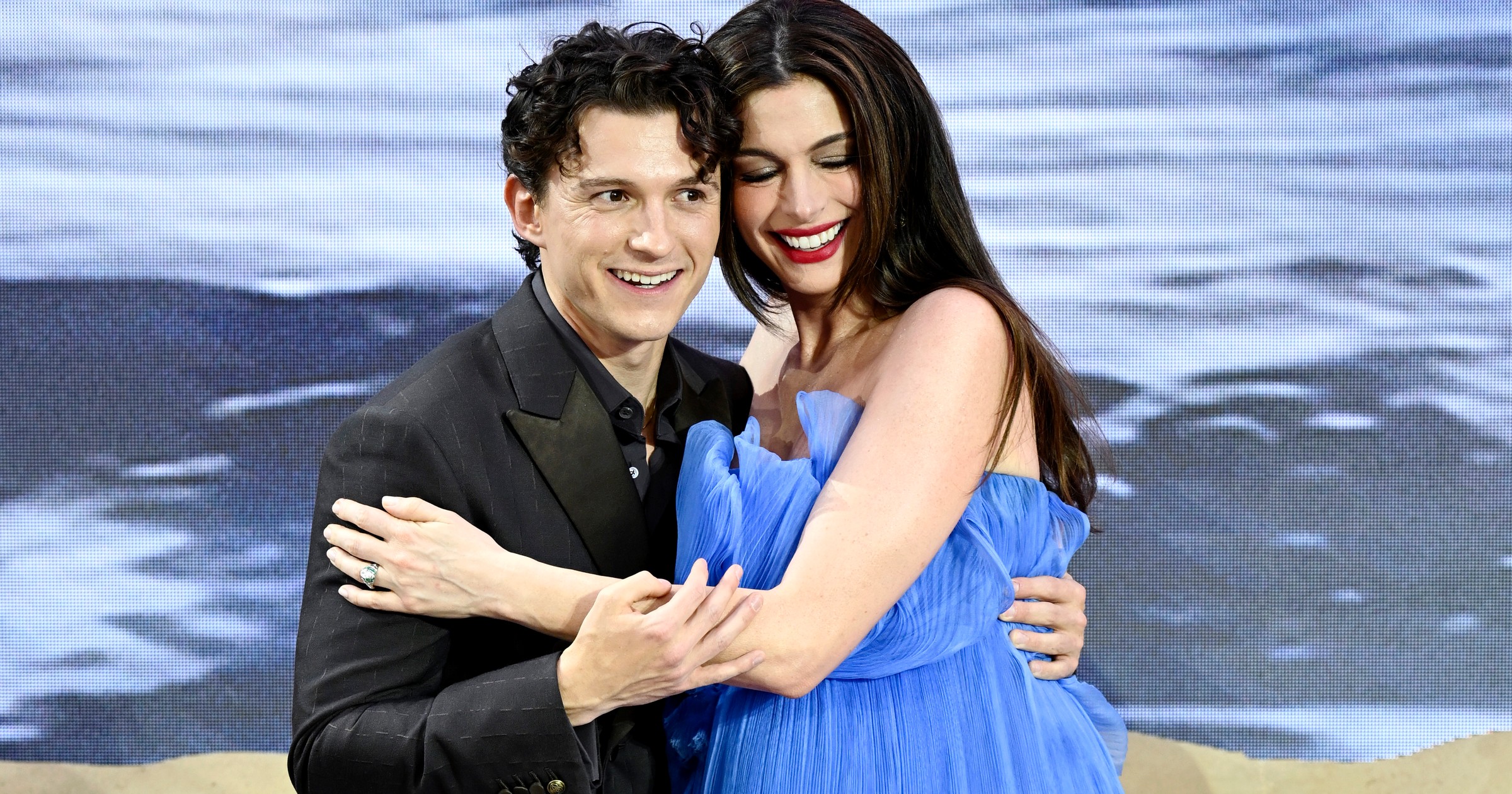 Tom Holland in a black suit and Anne Hathaway in a flowing blue strapless gown hugging and smiling at "The Odyssey" world premiere in London.