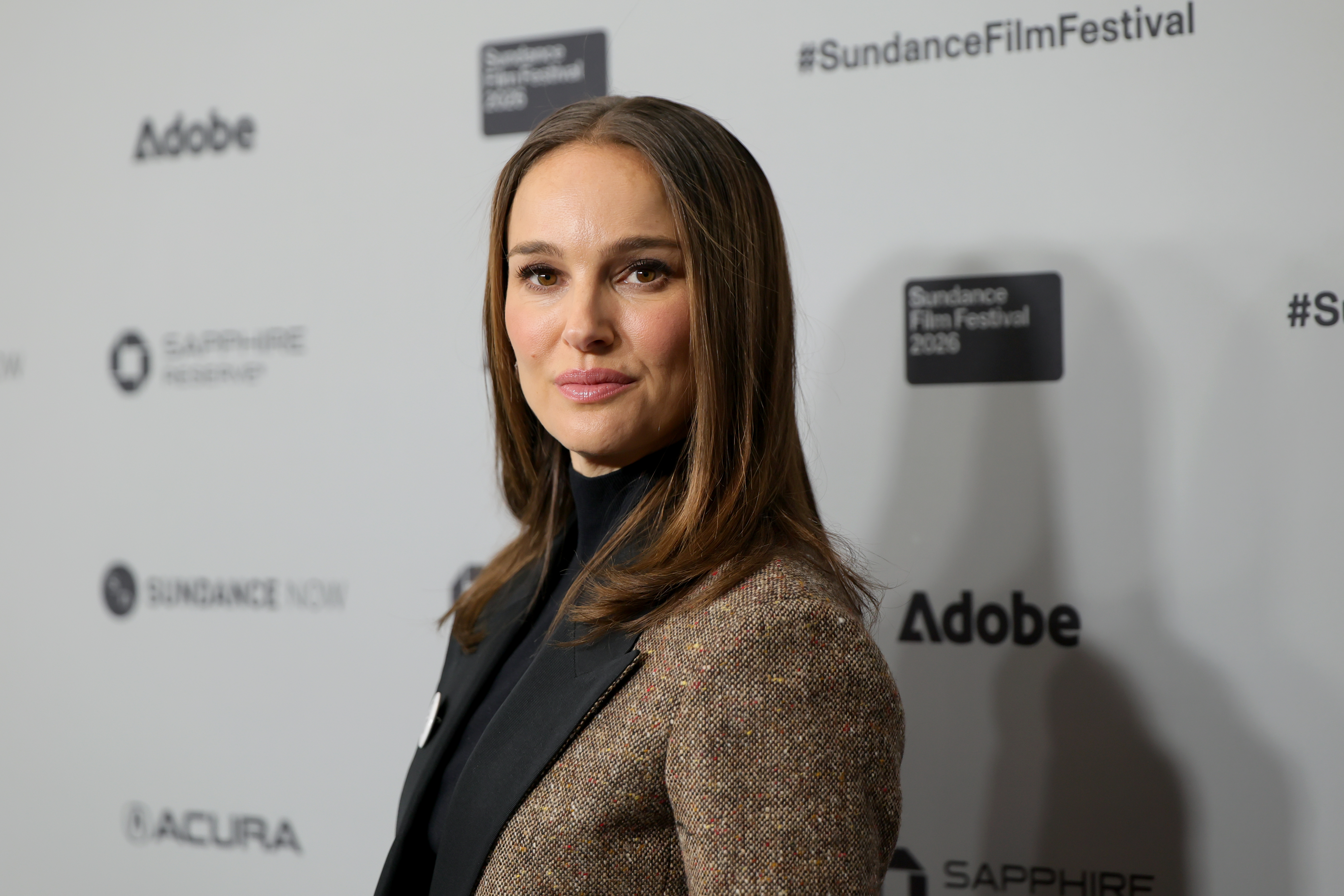 Natalie Portman in a tweed blazer and black turtleneck posing in front of a Sundance Film Festival step-and-repeat backdrop.