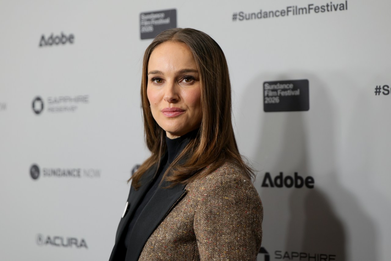 Natalie Portman in a tweed blazer and black turtleneck posing in front of a Sundance Film Festival step-and-repeat backdrop.