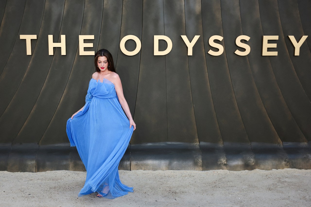 Anne Hathaway in a billowing blue strapless gown standing before a large gold "The Odyssey" sign at a premiere.