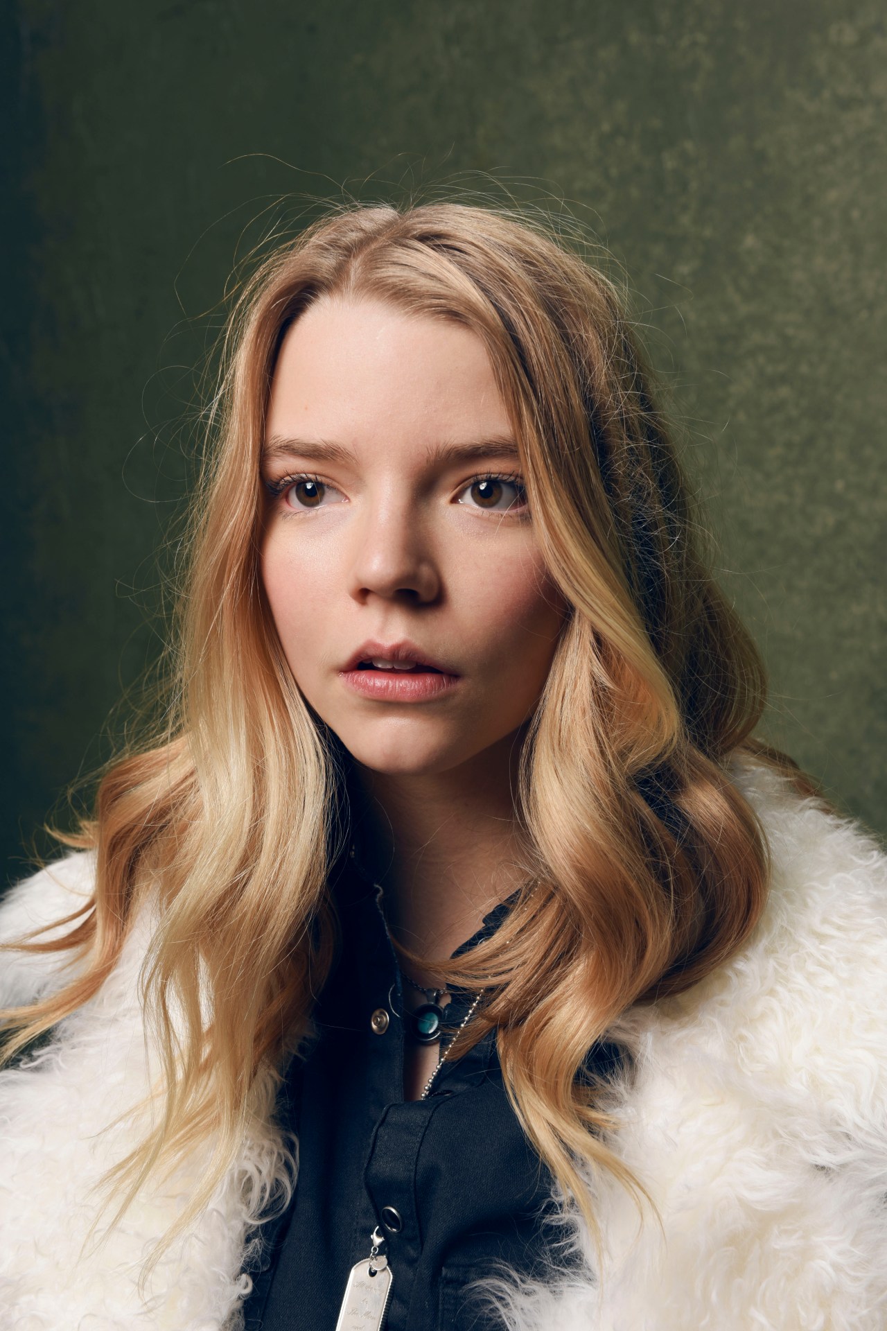 Young blonde woman with natural wavy hair wearing a white fur coat and dark top with a pendant necklace, photographed against a dark green background.