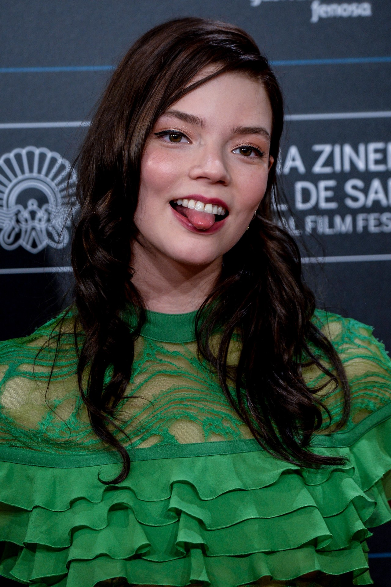 Dark-haired woman in a green lace ruffled top sticking her tongue out playfully at a film festival backdrop.