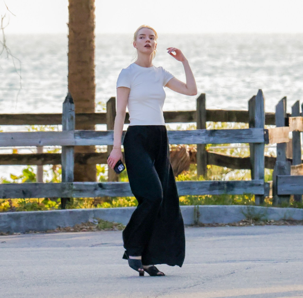 Blonde woman in a white fitted T-shirt and wide-leg black skirt walking along a paved path near a wooden fence by the water.