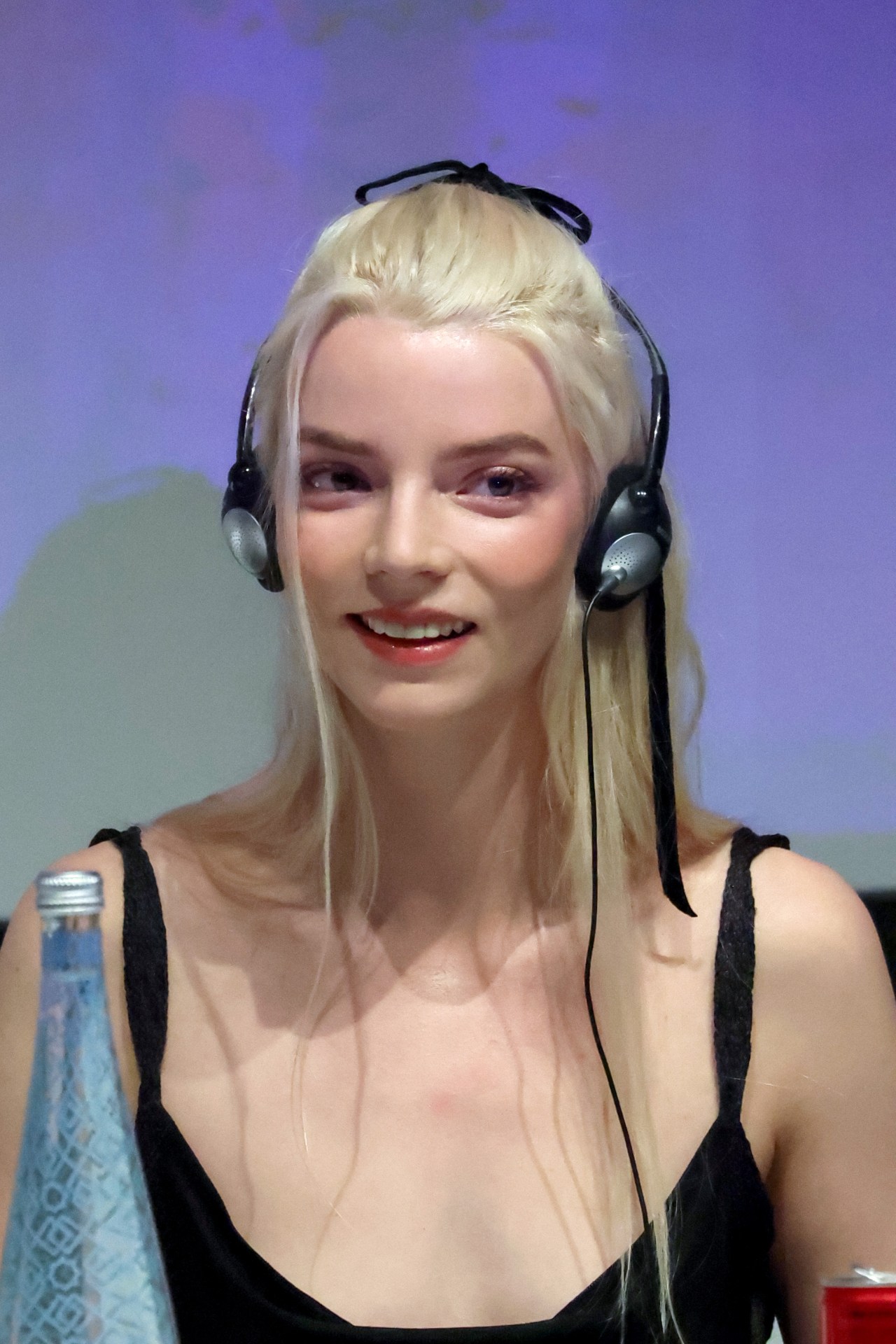 Blonde woman wearing black headphones and a black top, smiling slightly at a press conference table with a water bottle in the foreground.