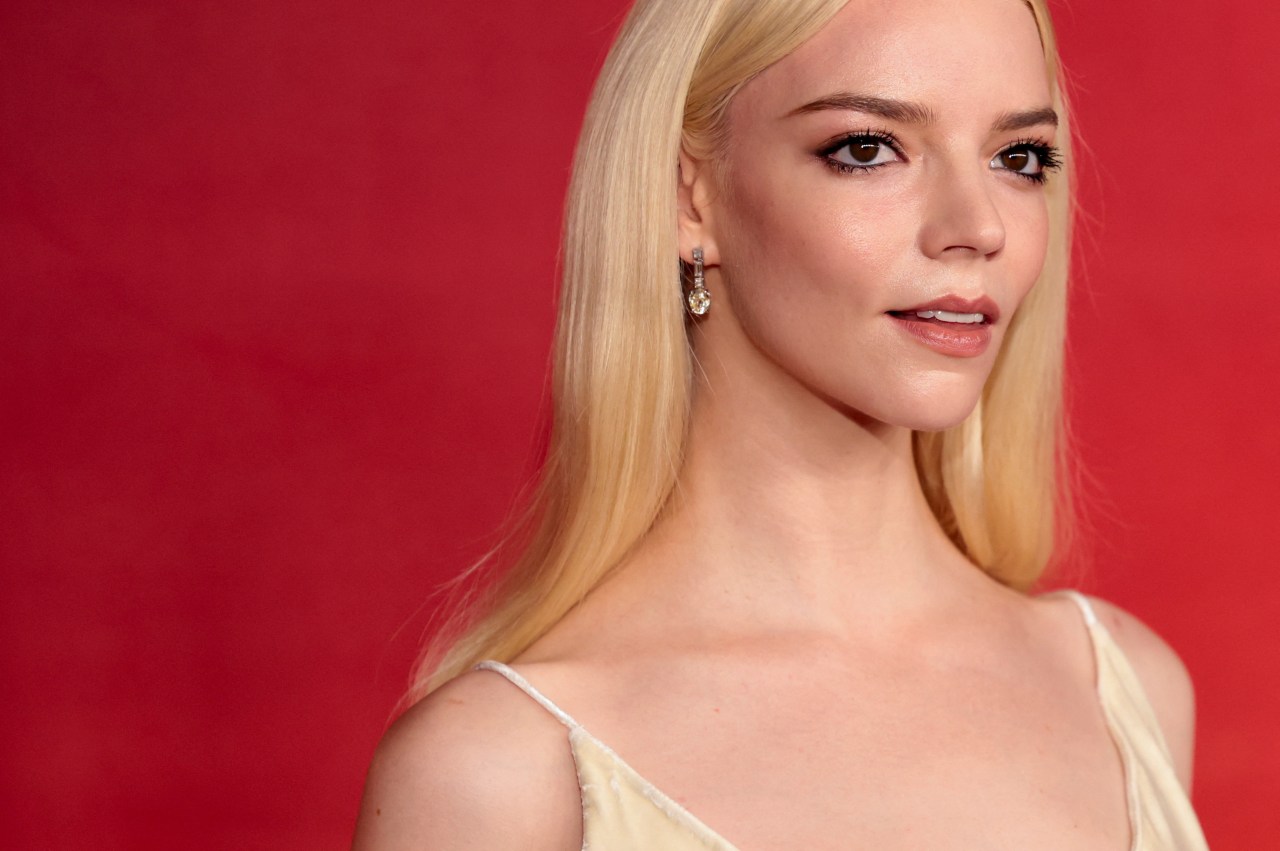 Close-up of a blonde woman in a white dress wearing a crystal drop earring, against a red background.