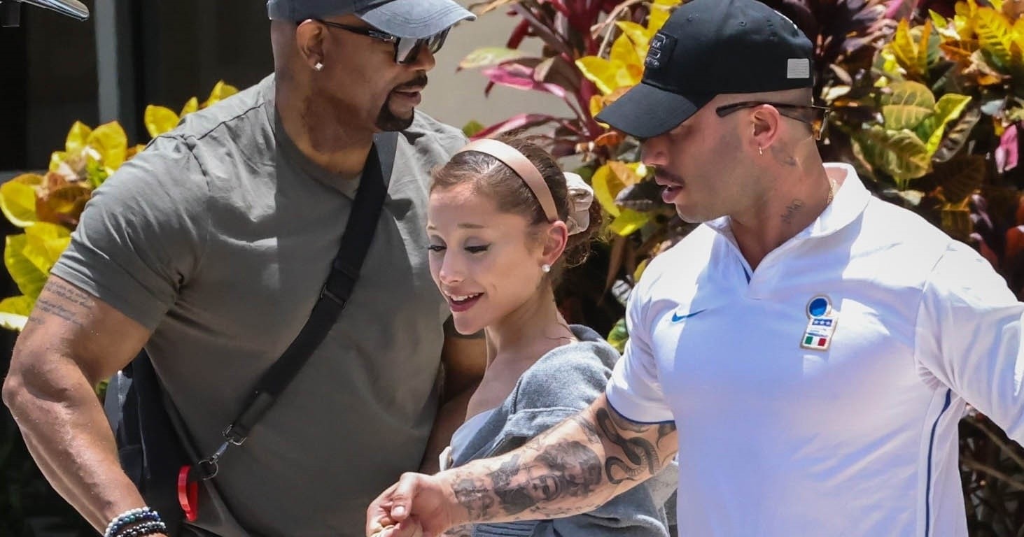 Ariana Grande walking out of Whole Foods carrying brown paper bags, with two men on either side pushing a shopping cart.