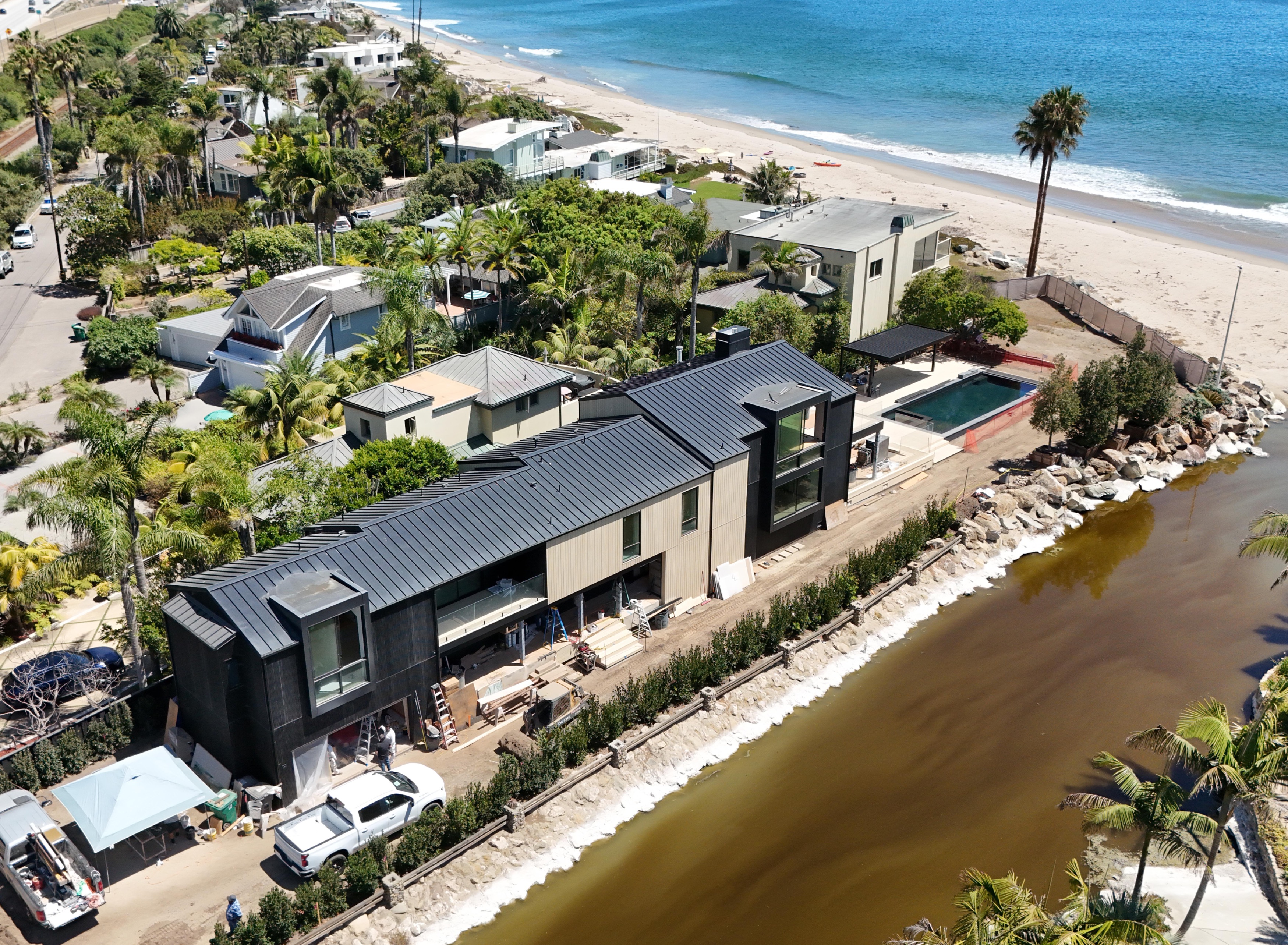 Aerial view of a modern dark-roofed beachfront estate under construction beside a lagoon, with a lap pool and the Pacific Ocean visible in the background.