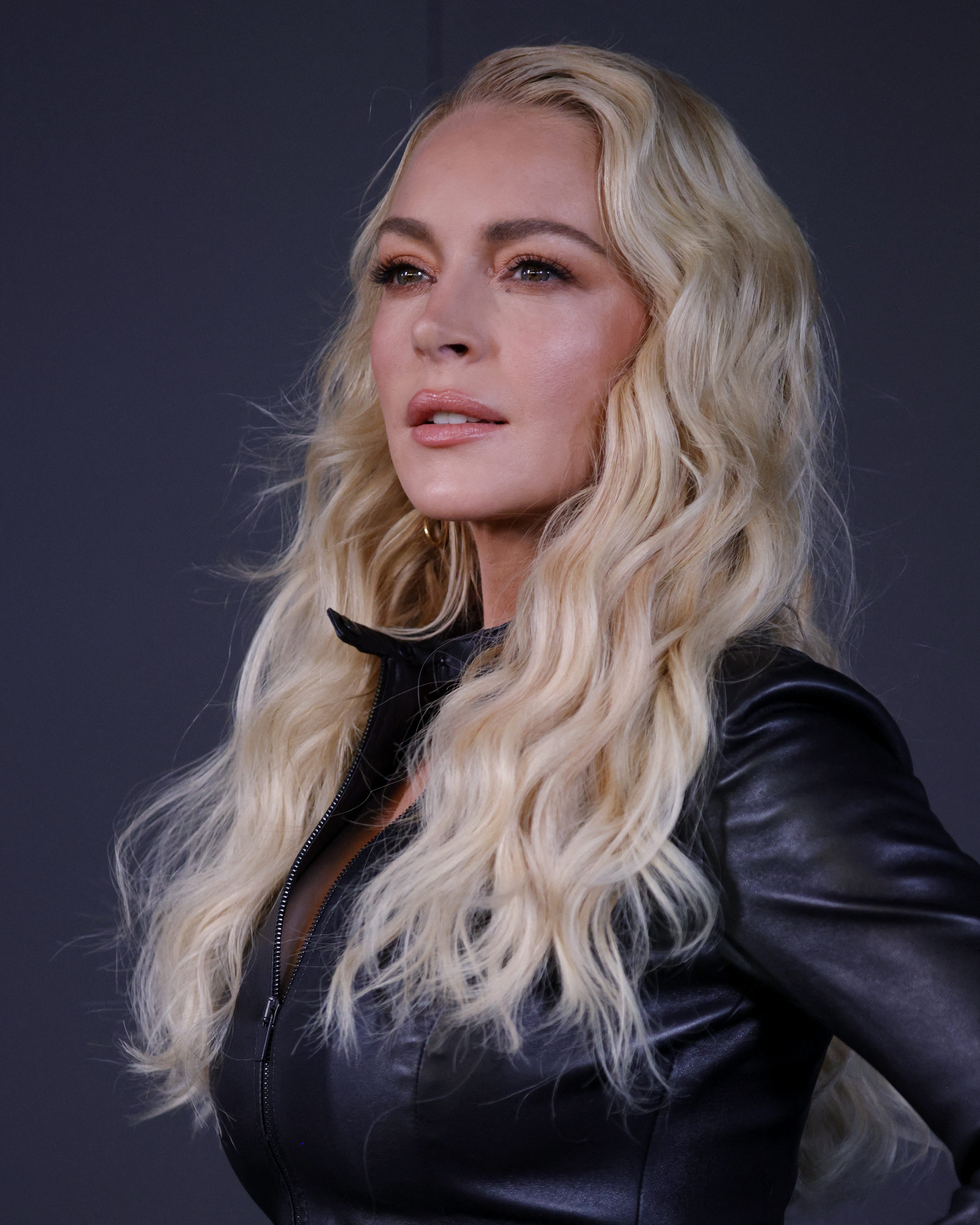 Click to open the image gallery Woman with long blonde wavy hair wearing a black leather zip-up jacket, gazing upward against a dark background.