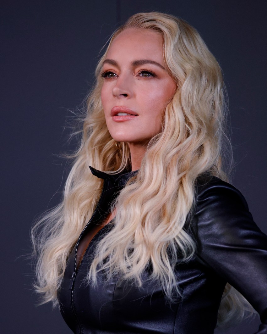 Woman with long blonde wavy hair wearing a black leather zip-up jacket, gazing upward against a dark background.