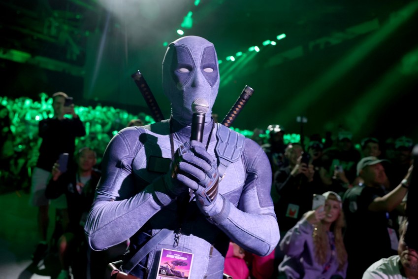 Person in a grey Deadpool costume holding a microphone at a large arena event with green lighting and a crowd in the background.