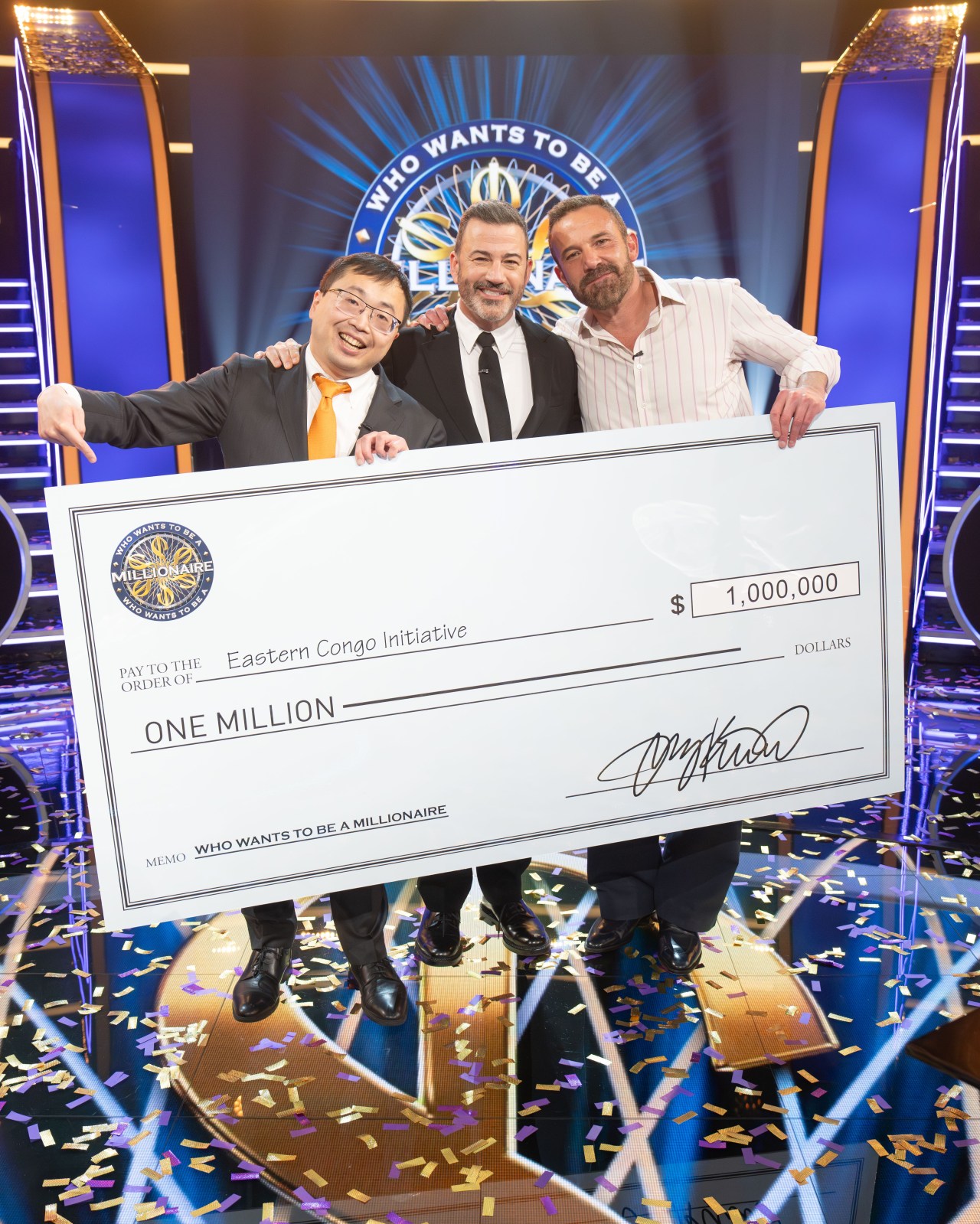 Three men hold a giant $1 million check made out to Eastern Congo Initiative on the Who Wants to Be a Millionaire stage, surrounded by falling confetti.