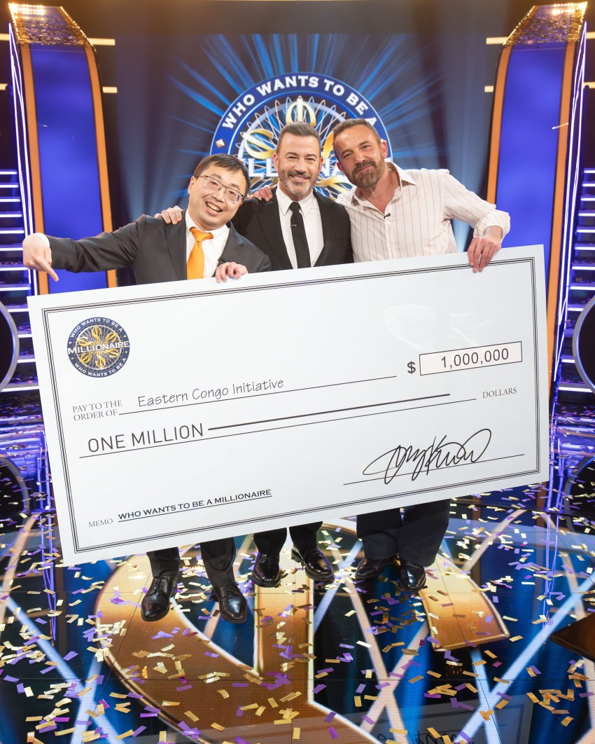 Three men hold a giant $1 million check made out to Eastern Congo Initiative on the Who Wants to Be a Millionaire stage, surrounded by falling confetti.