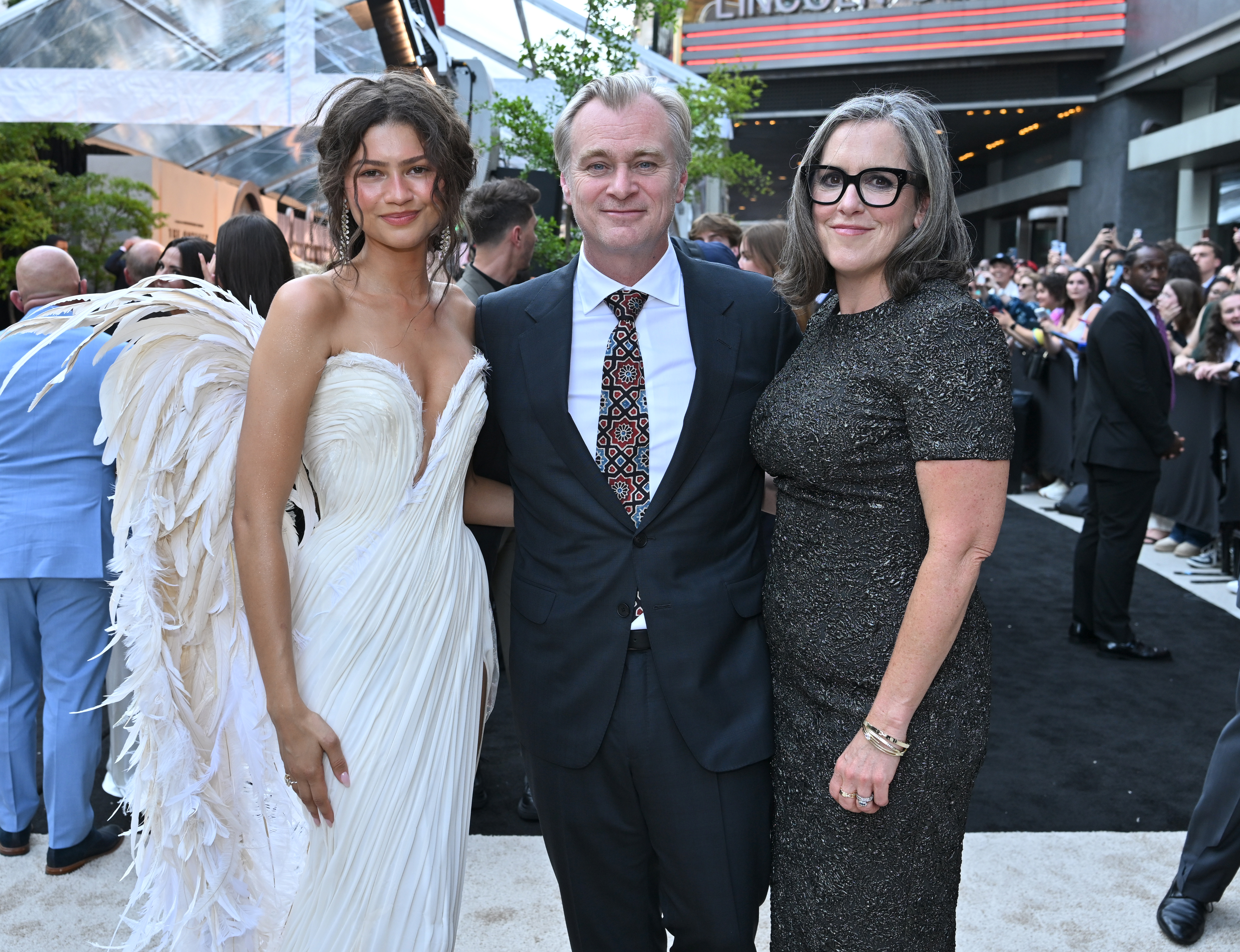 Zendaya in a white feathered gown, Christopher Nolan in a black suit, and Emma Thomas in a dark beaded dress pose together at an outdoor premiere event.