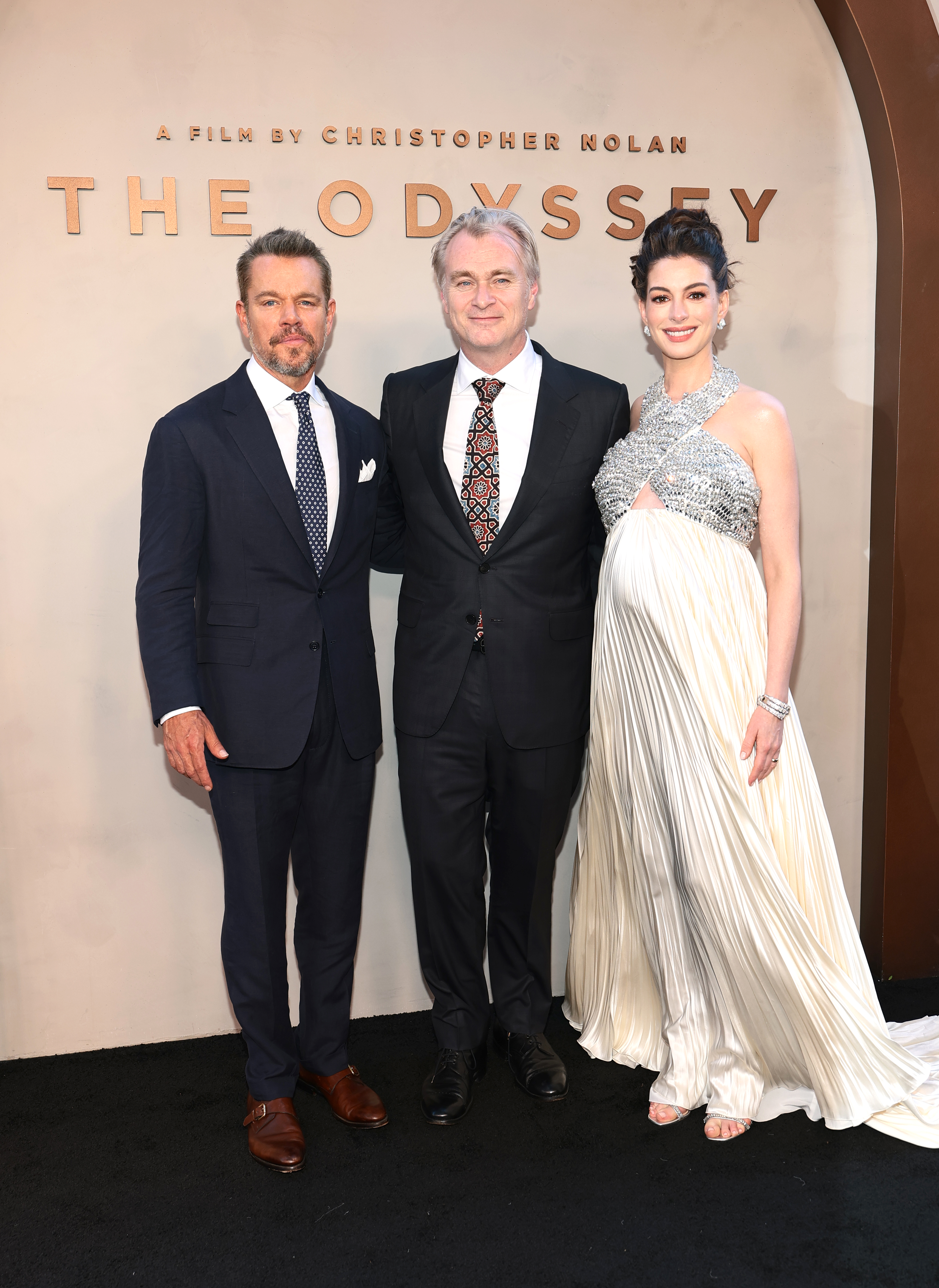Matt Damon in a navy suit, Christopher Nolan in a black suit, and Anne Hathaway in a silver and white pleated gown stand in front of 'The Odyssey' premiere backdrop.