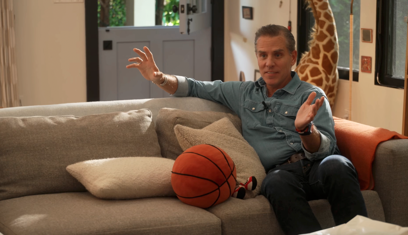 Silver-haired man in a denim shirt seated on a couch, gesturing with both hands, a plush basketball beside him and a giant stuffed giraffe in the background.