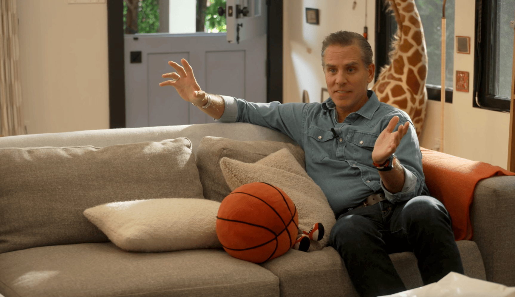 Silver-haired man in a denim shirt seated on a couch, gesturing with both hands, a plush basketball beside him and a giant stuffed giraffe in the background.