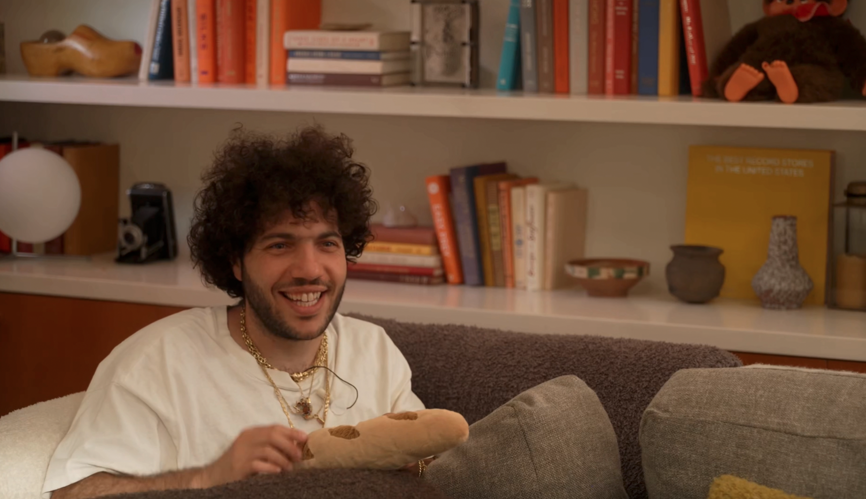 Curly-haired young man in a white t-shirt and gold chains laughing on a couch, holding a stuffed plush baguette, bookshelves in background.