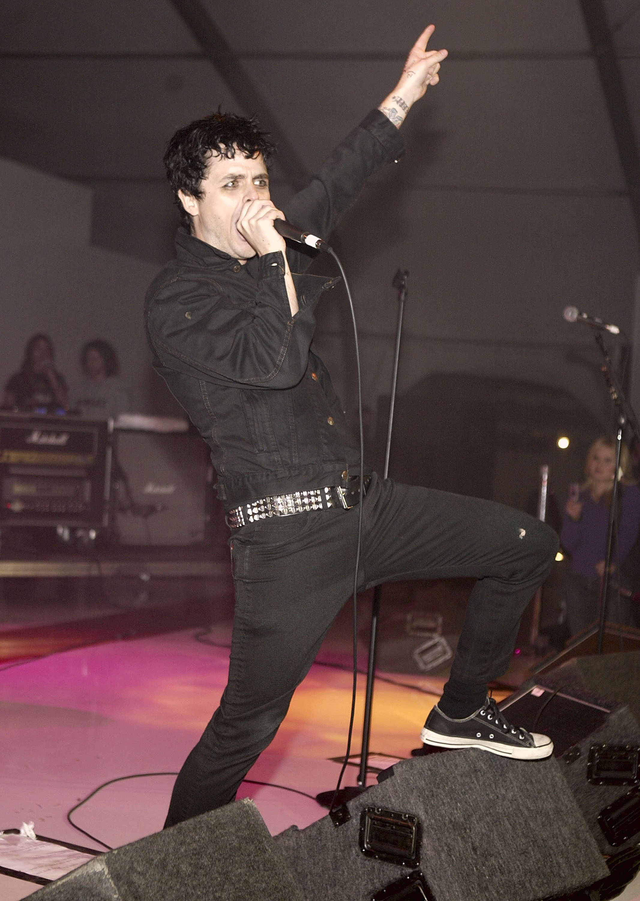Billie Joe Armstrong singing into a microphone with one arm raised on stage, wearing all black with a studded belt and Converse sneakers.