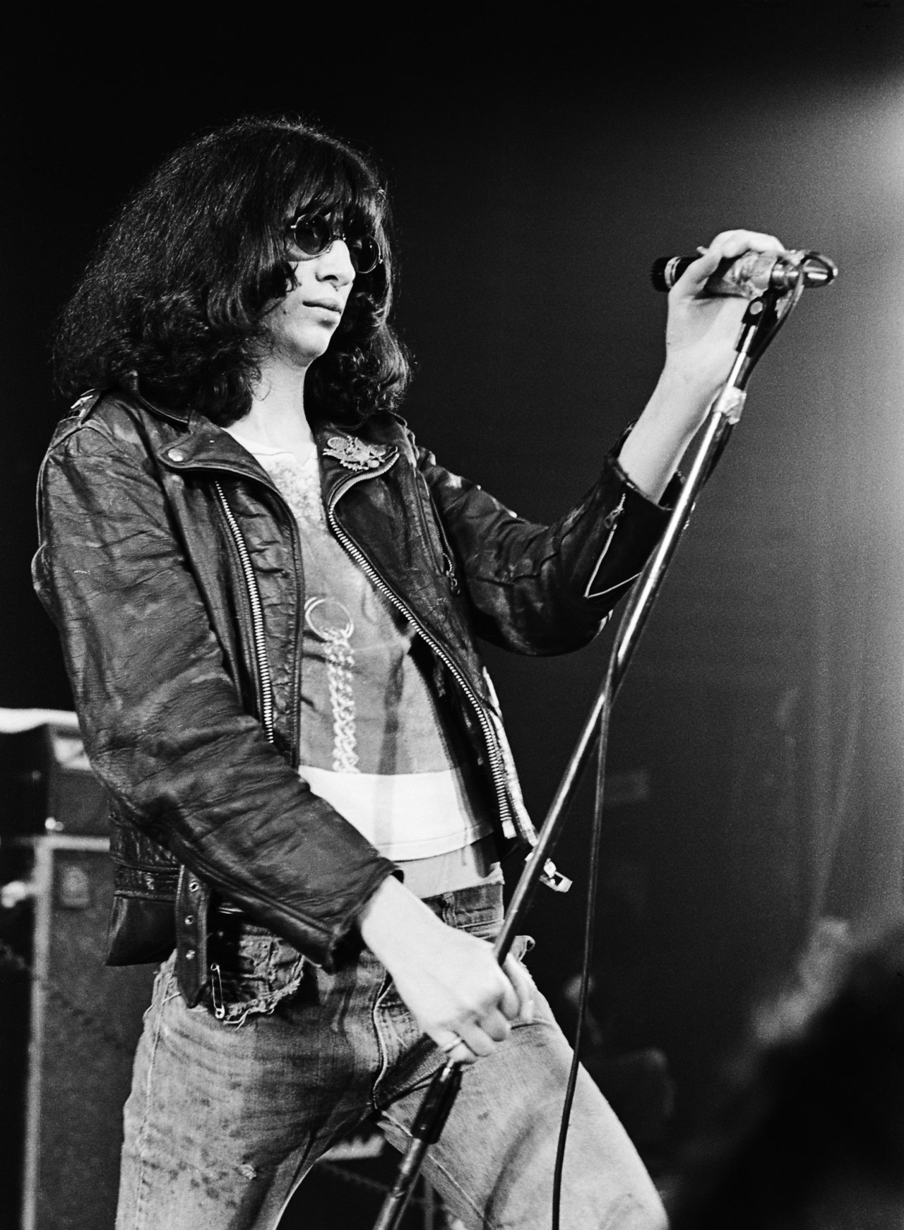Black-and-white photo of a long-haired male singer in a leather jacket and sunglasses gripping a microphone stand on stage.